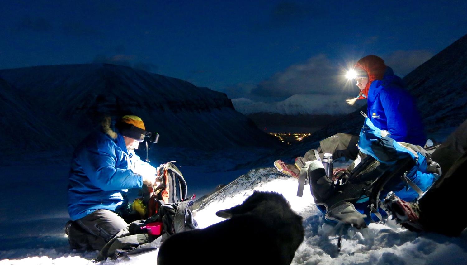 Two people with headlamps and a dog in the foreground, with mountains and Longyearbyen in the background.
