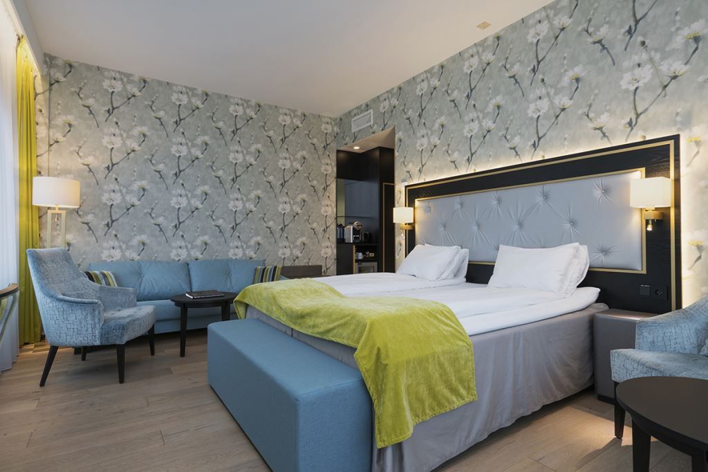 Interior of hotel room: Double bed, blue armchair and sofa, floral wallpaper.