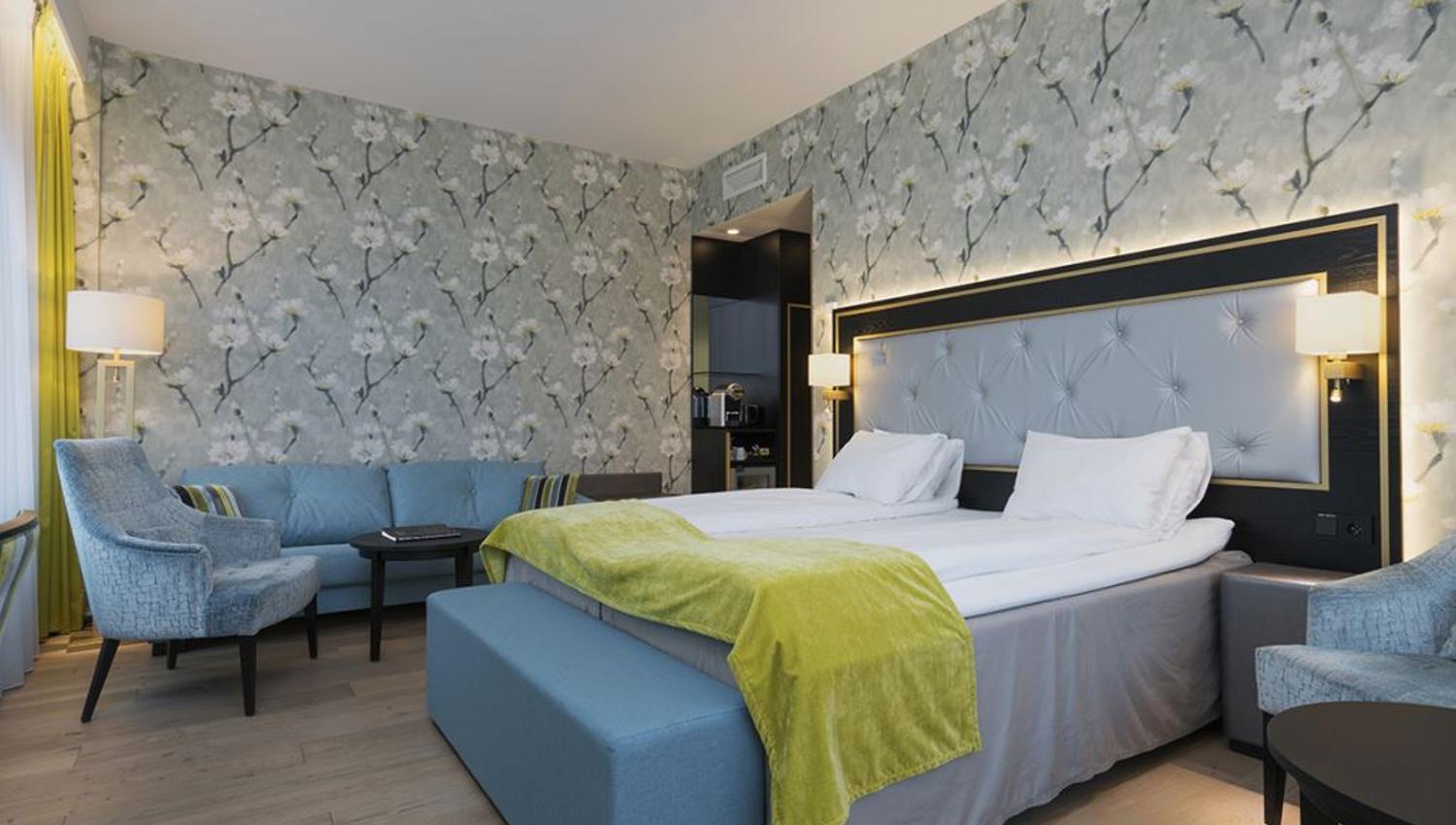 Interior of hotel room: Double bed, blue armchair and sofa, floral wallpaper.