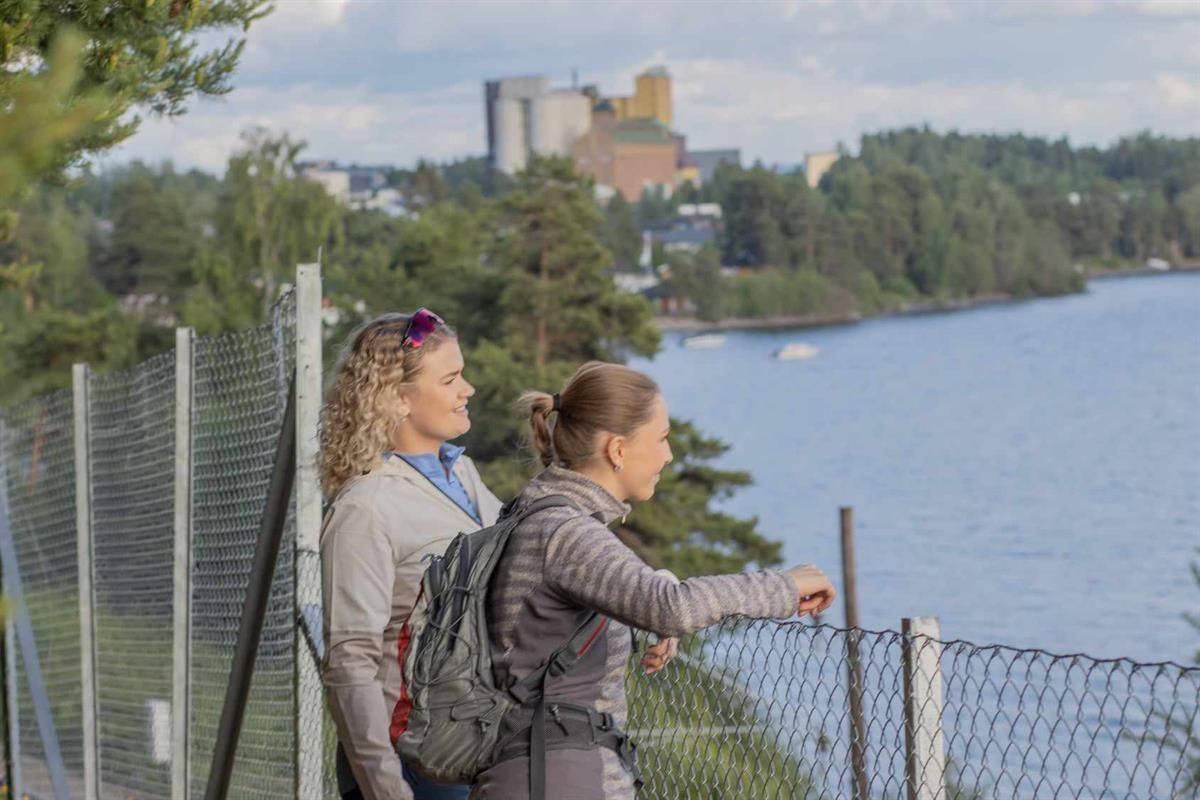 To personer står ved et gjerde og ser utover en innsjø og bygninger på den andre siden.