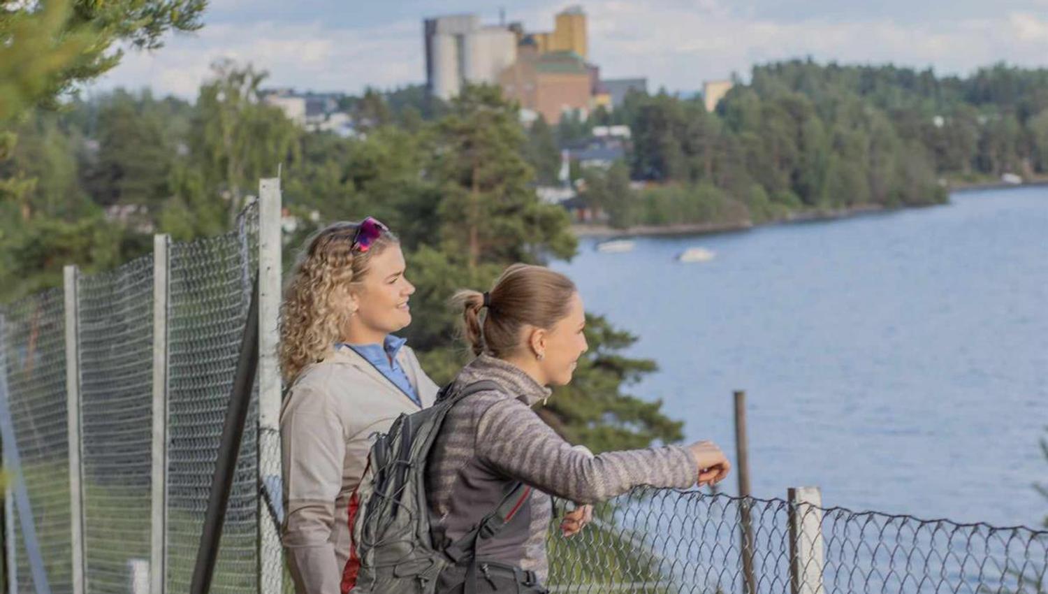 To personer står ved et gjerde og ser utover en innsjø og bygninger på den andre siden.