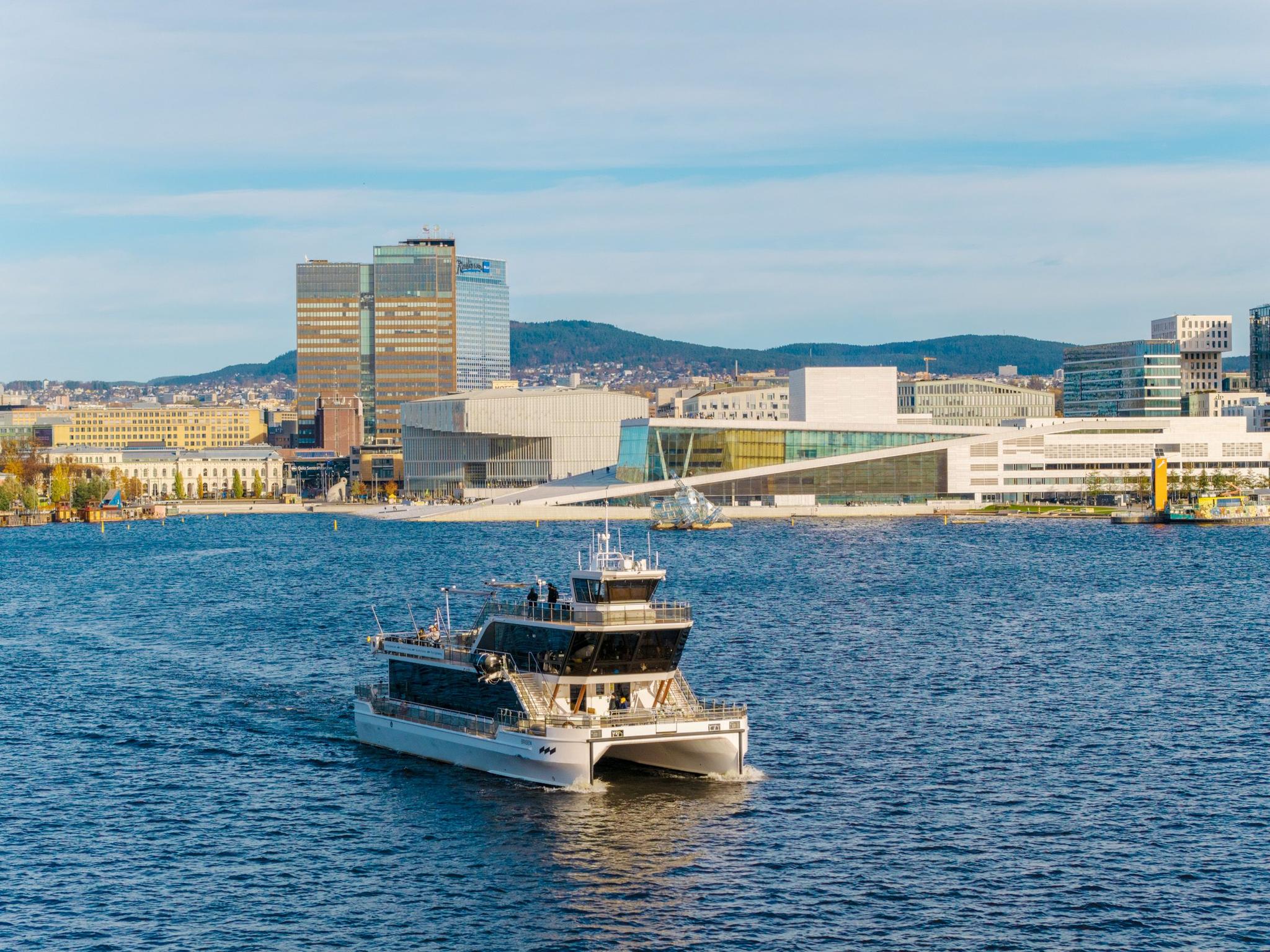 Dronebilde av båten i fjorden med byen i bakgrunnen