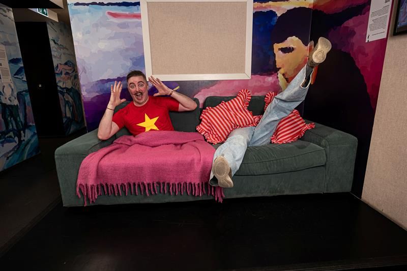 En person ligger leende i en sofa i et fargerikt museumsrom med beinene strukket opp og hendene i været.