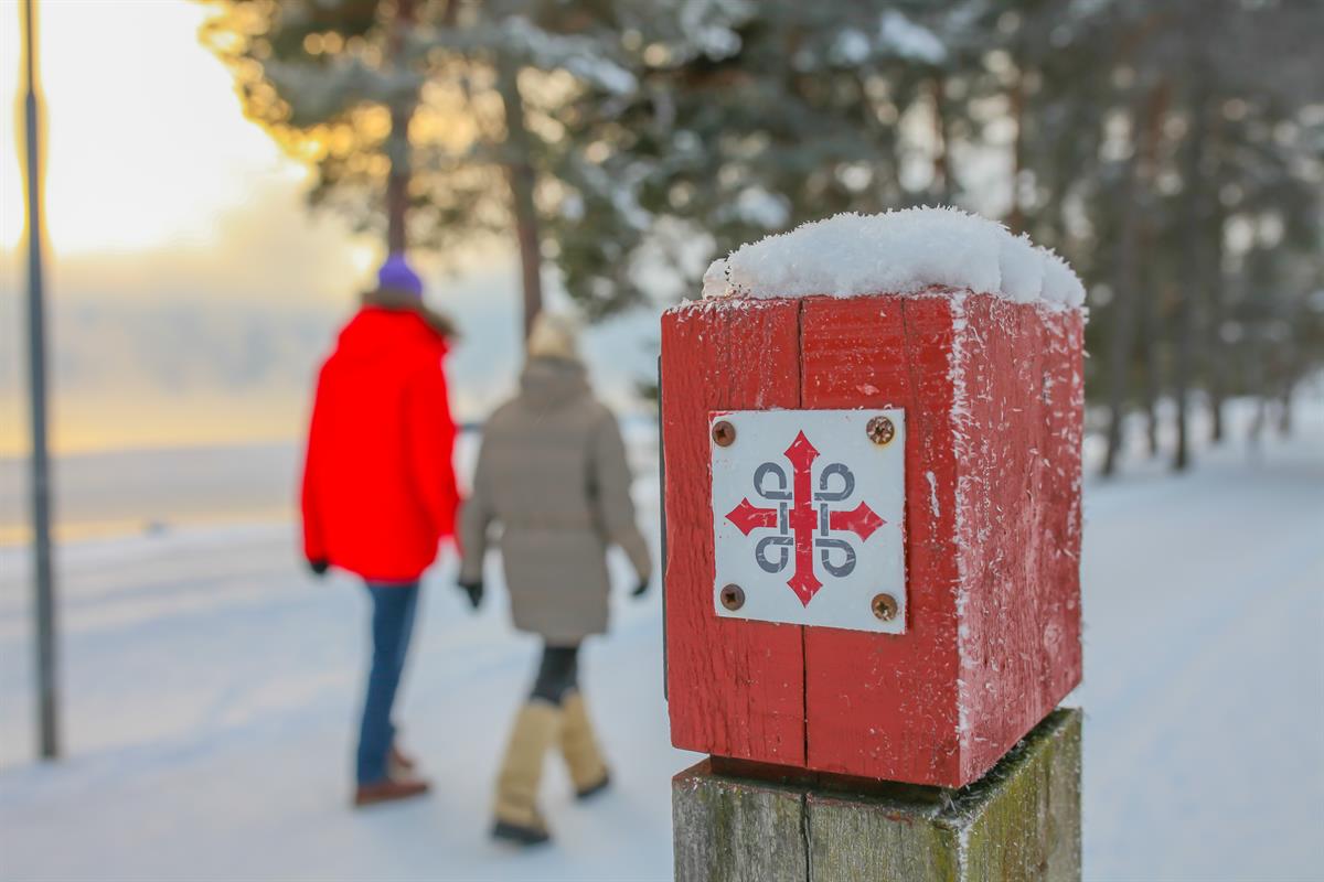 En rød merkestolpe med et hvitt symbol står i snøen, med to personer som går i bakgrunnen.