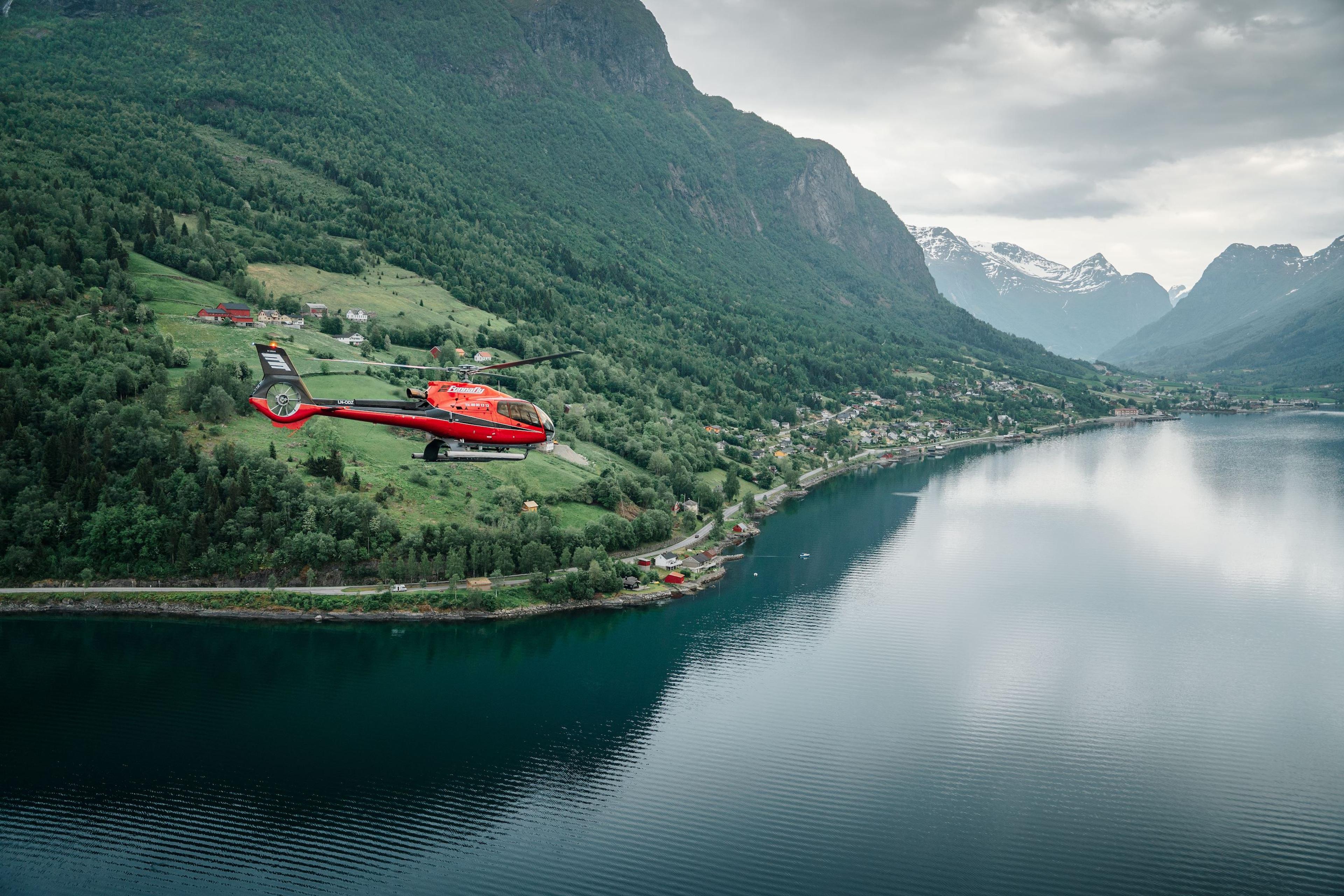 Fjord Helikopter - A red helicopter flies above a calm fjord with forested mountains, small villages, and cloudy skies in the distance..