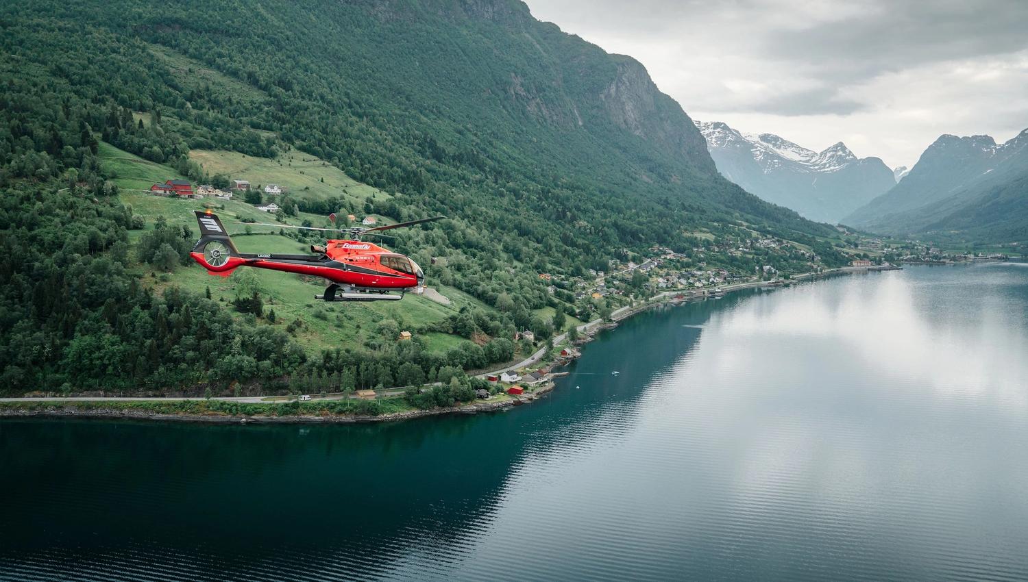 Fjord Helikopter - Eit raudt helikopter flyg over ein fjord med skogkledde fjell, bygder langs stranda og skya himmel..