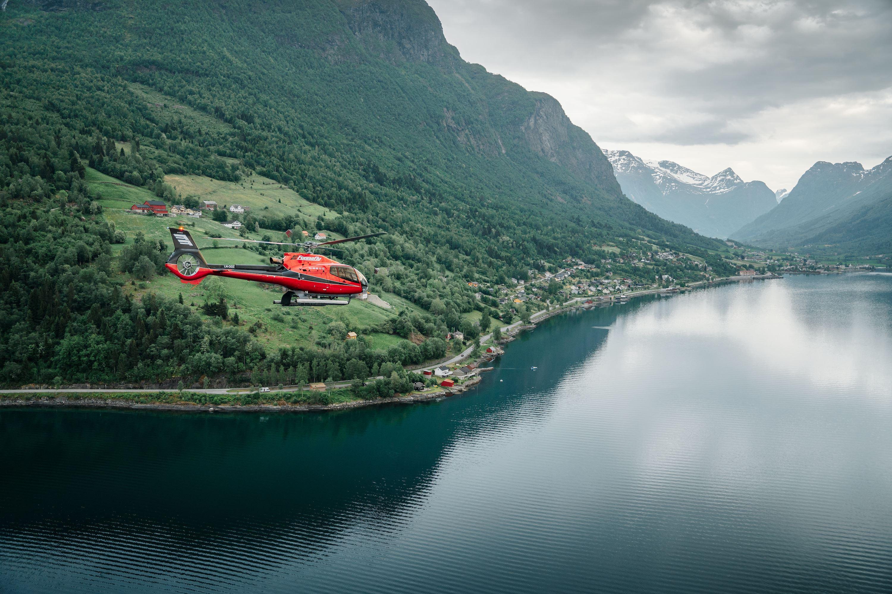 Fjord Helikopter - A red helicopter flies above a calm fjord with forested mountains, small villages, and cloudy skies in the distance..