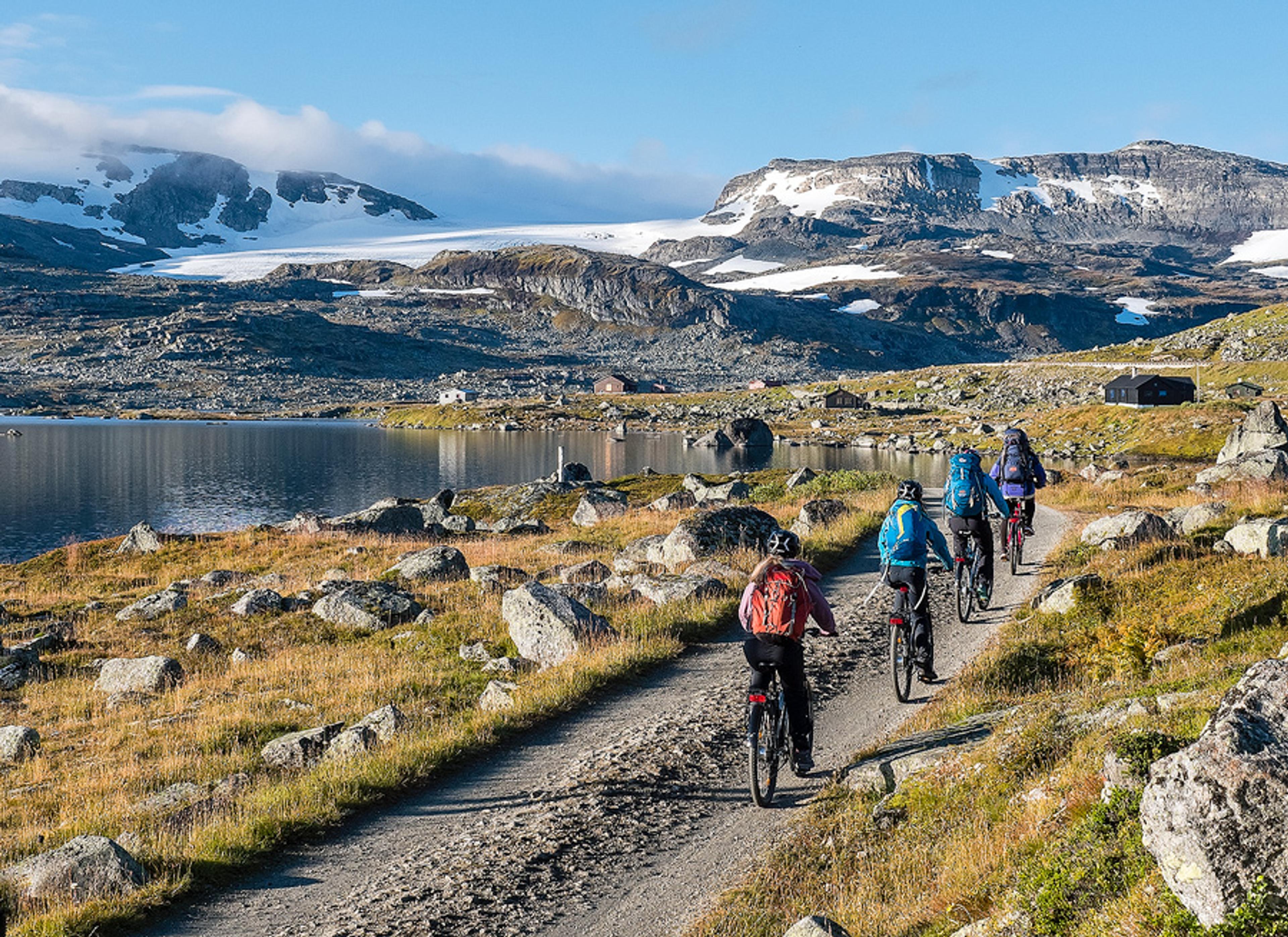 Cyclists on the Rallarvegen route in Hardanger, surrounded by snowy mountains and glaciers on a clear summer day.