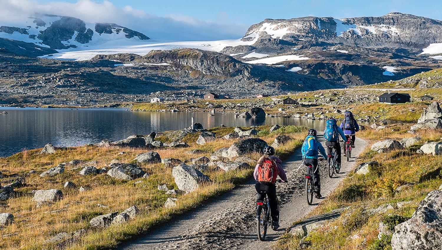 Syklistar på Rallarvegen i Hardanger med utsikt mot snødekte fjell og brear under ein klår sommarhimmel.