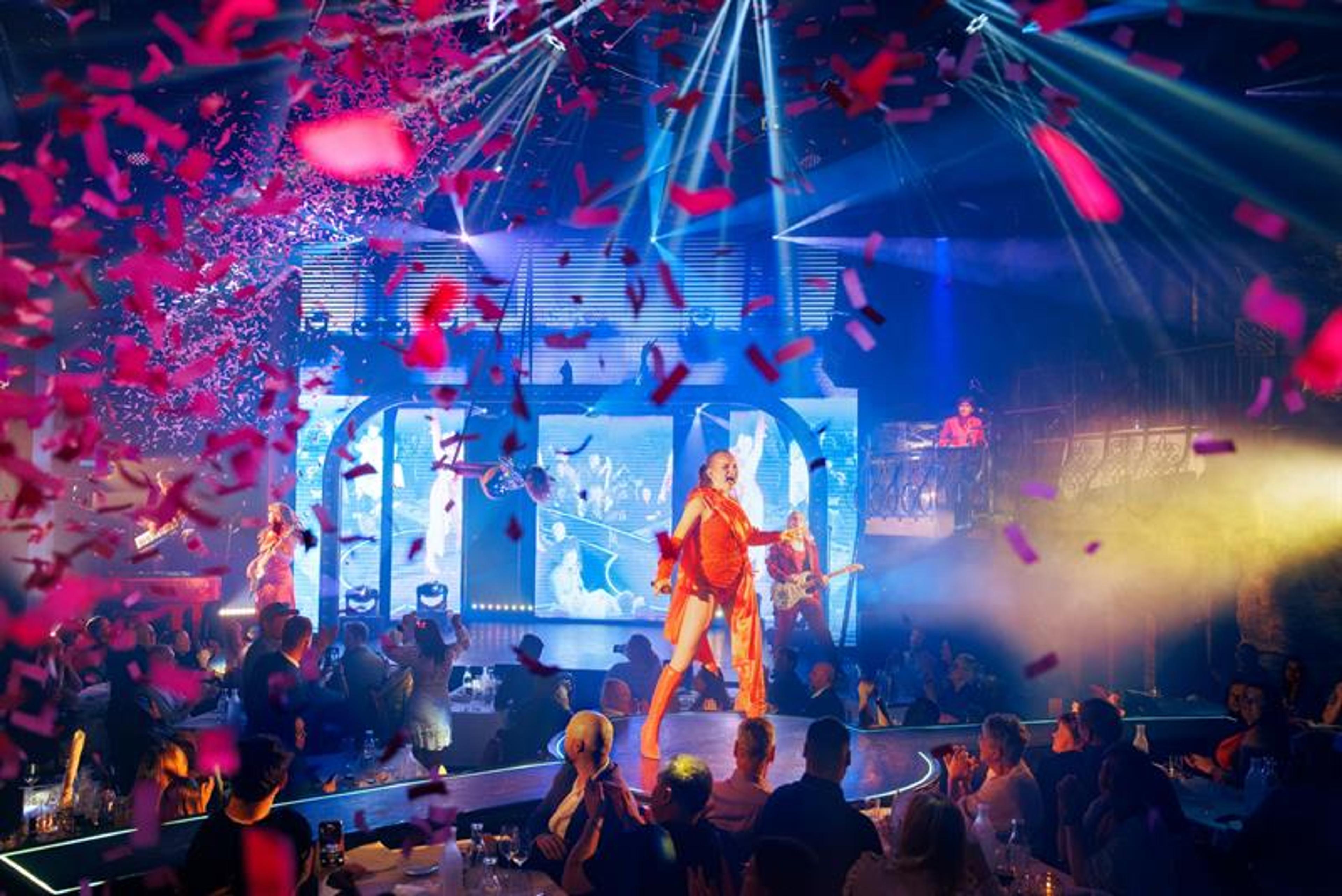 A performer in red leads the show as confetti rains down and the crowd cheers. Colourful lights and energy fill the room.