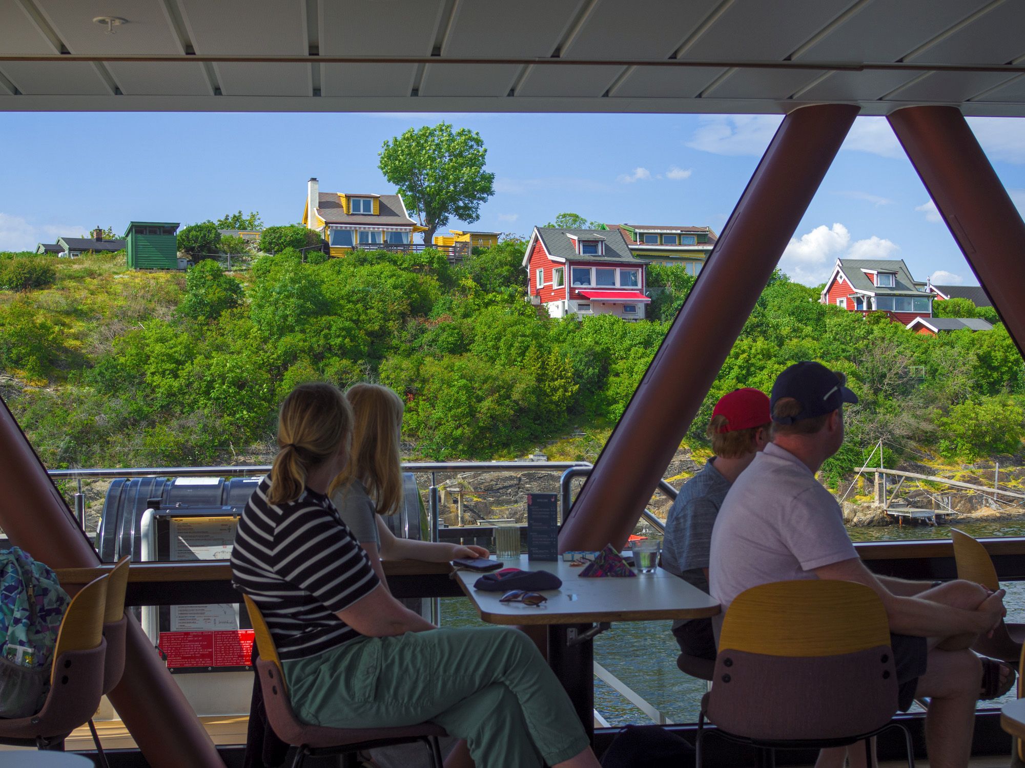 People sitting inside a boat looking out through the panoramic window at colourful houses and green landscape on the shore.