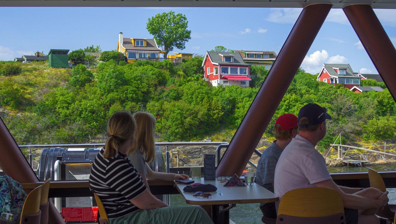 People sitting inside a boat looking out through the panoramic window at colourful houses and green landscape on the shore.