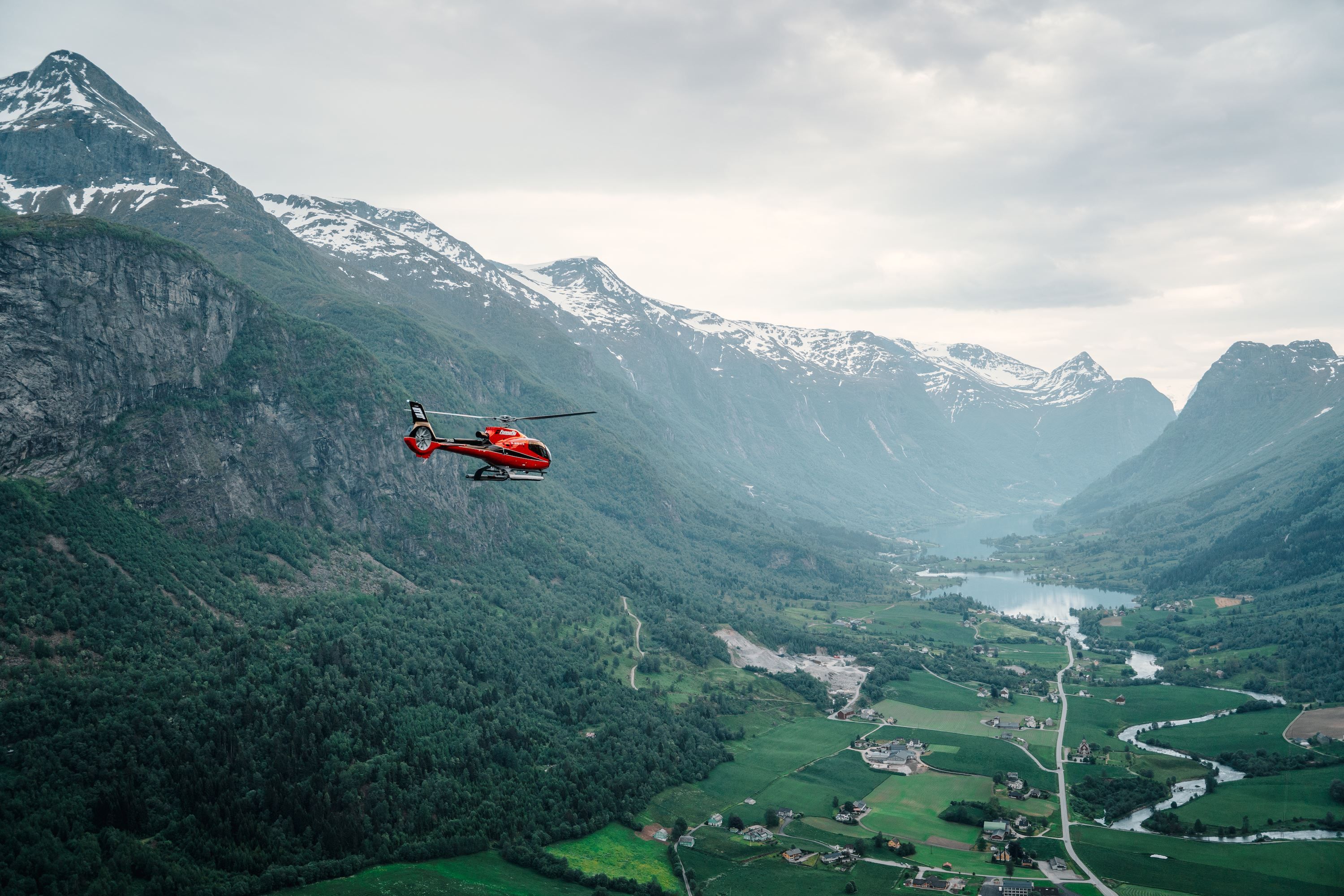 Fjord Helikopter - Eit raudt helikopter flyg over ei grøn fjelldal med snødekte fjell, elv, gardar og ein innsjø under skydekke..