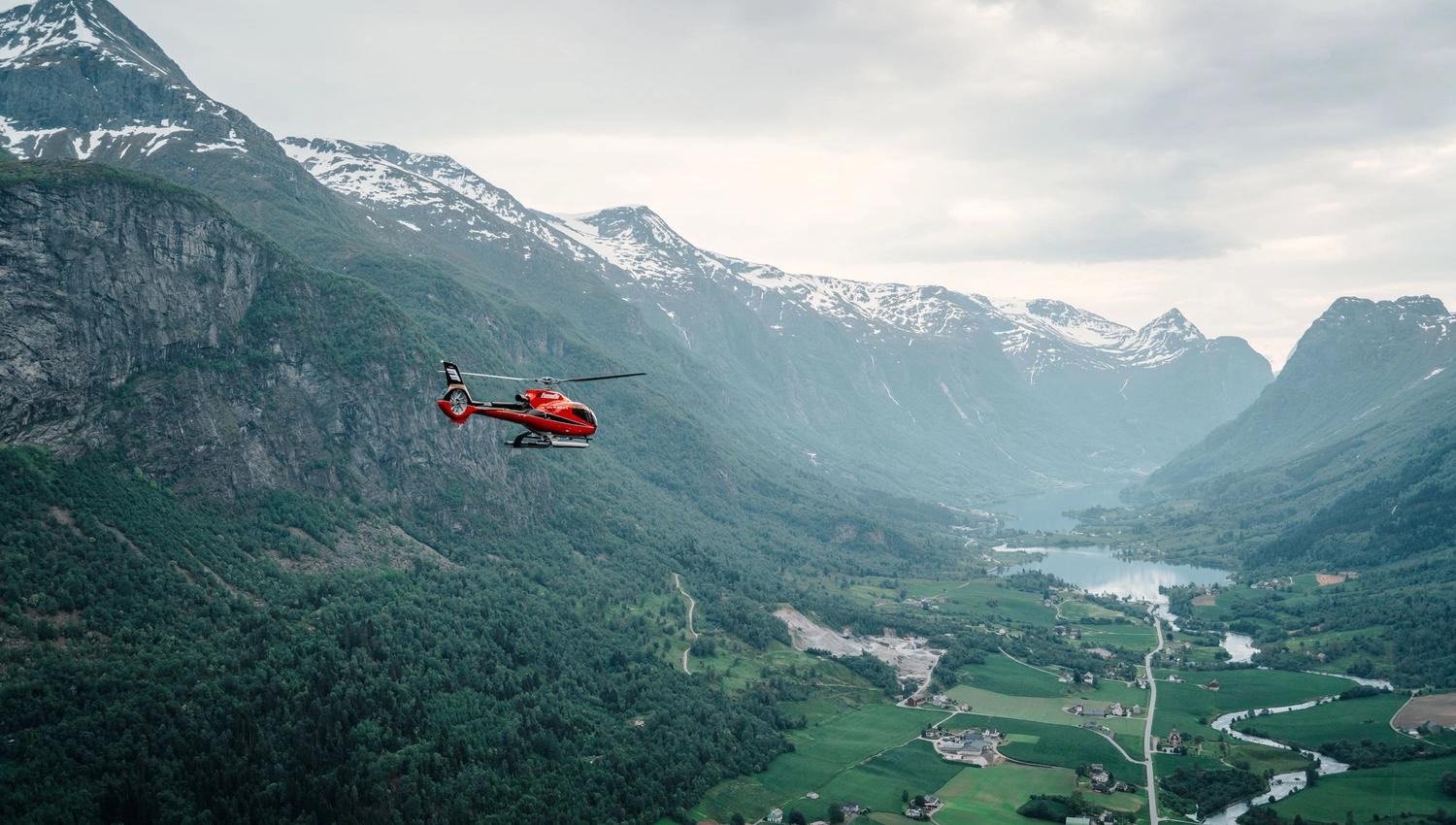 Fjord Helikopter - Eit raudt helikopter flyg over ei grøn fjelldal med snødekte fjell, elv, gardar og ein innsjø under skydekke..