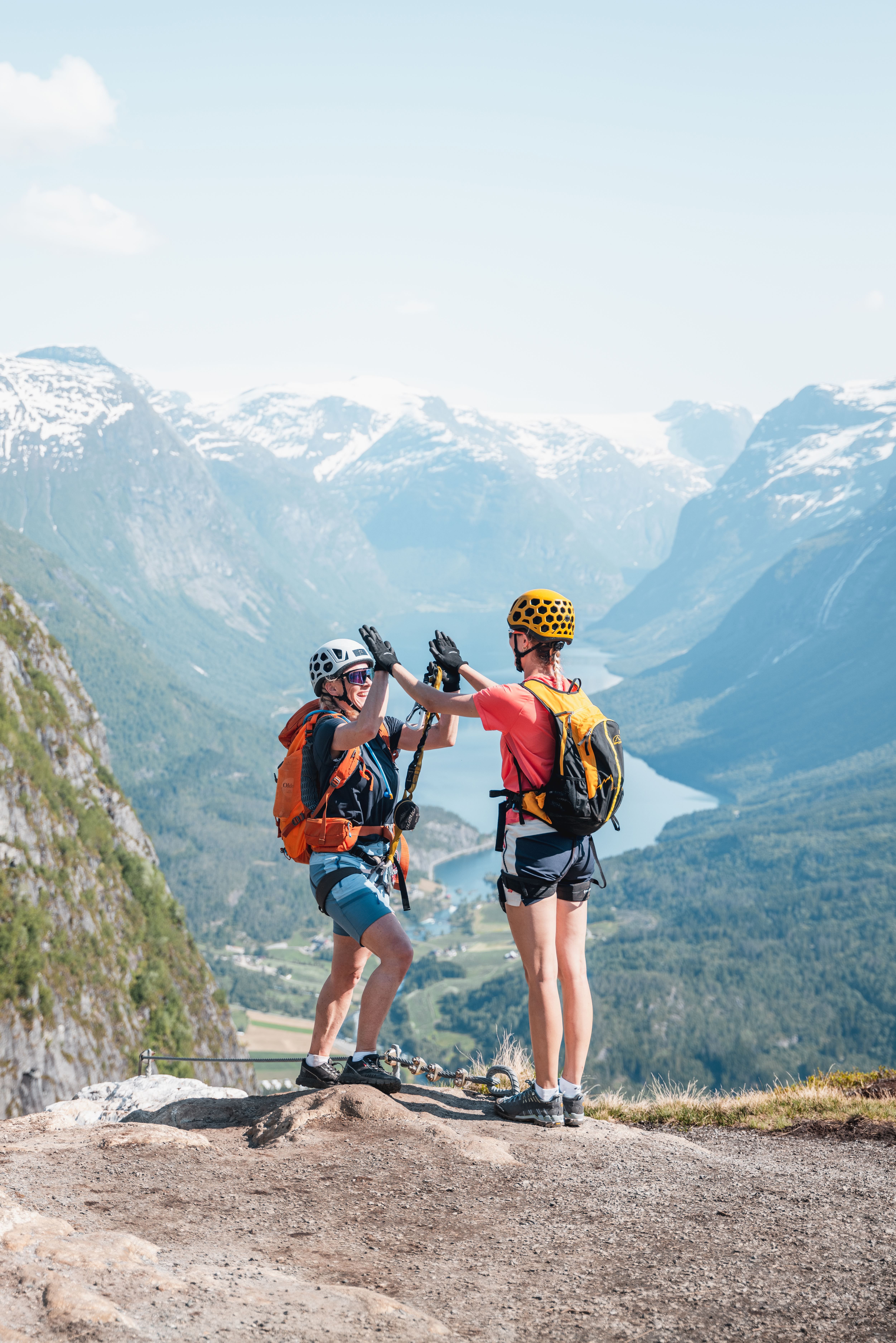 To klatrarar med hjelm og ryggsekk gir kvarandre høgfem på ein fjellkant med fjord og snødekte fjell i bakgrunnen..