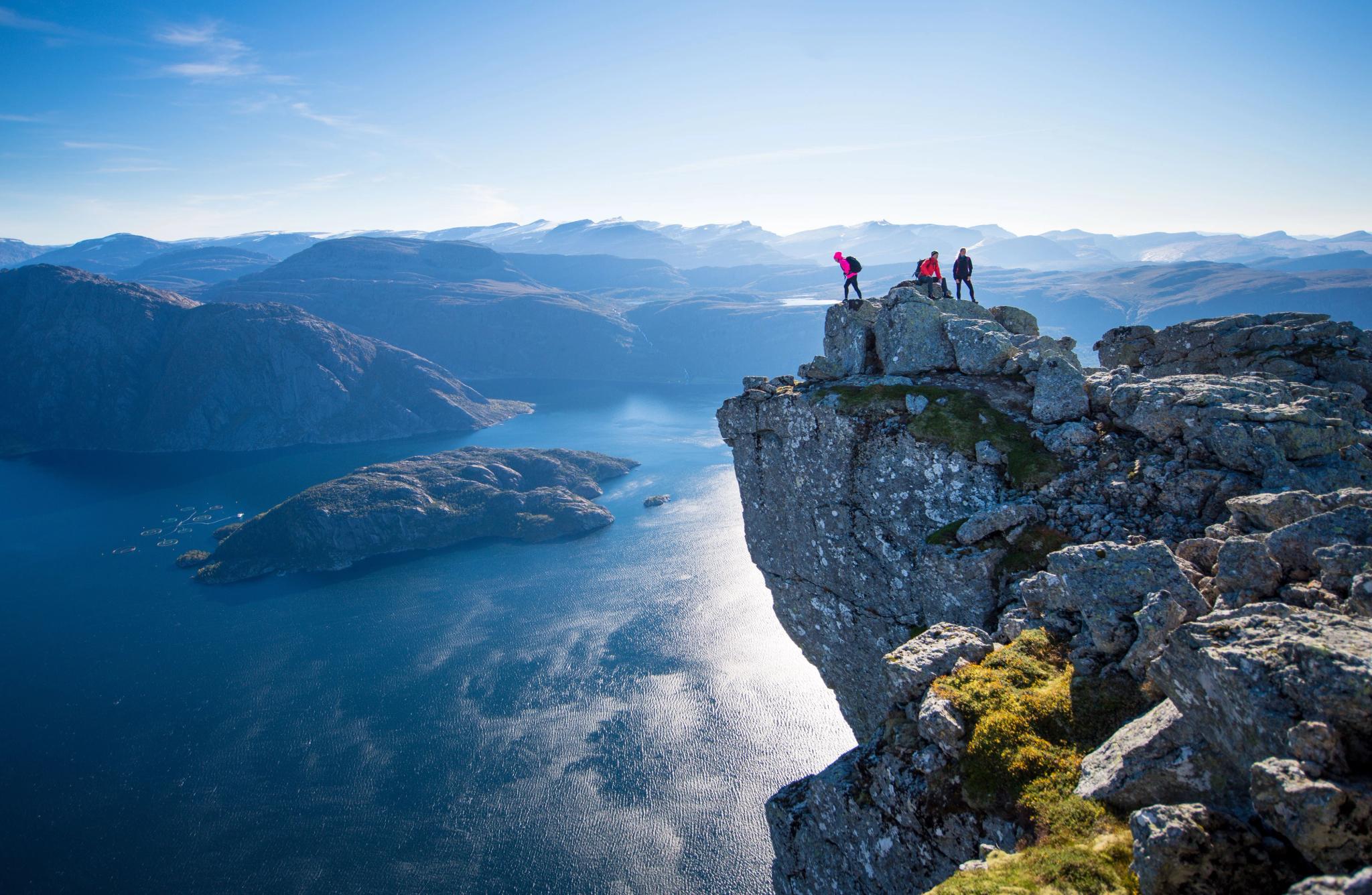 Three hikers stand on a rocky cliff edge overlooking a blue fjord and distant mountain ridges under clear sky..