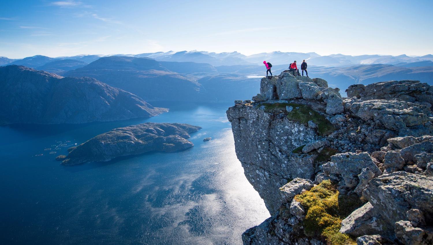 Three hikers stand on a rocky cliff edge overlooking a blue fjord and distant mountain ridges under clear sky..