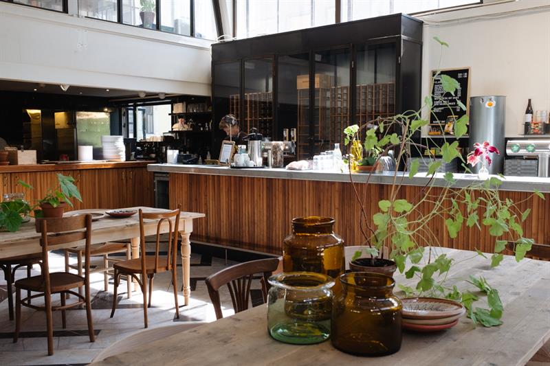 Two solid wood square dining tables with various glass jars and green plants on them, wooden chairs around the tables, a long bar counter, and a staff