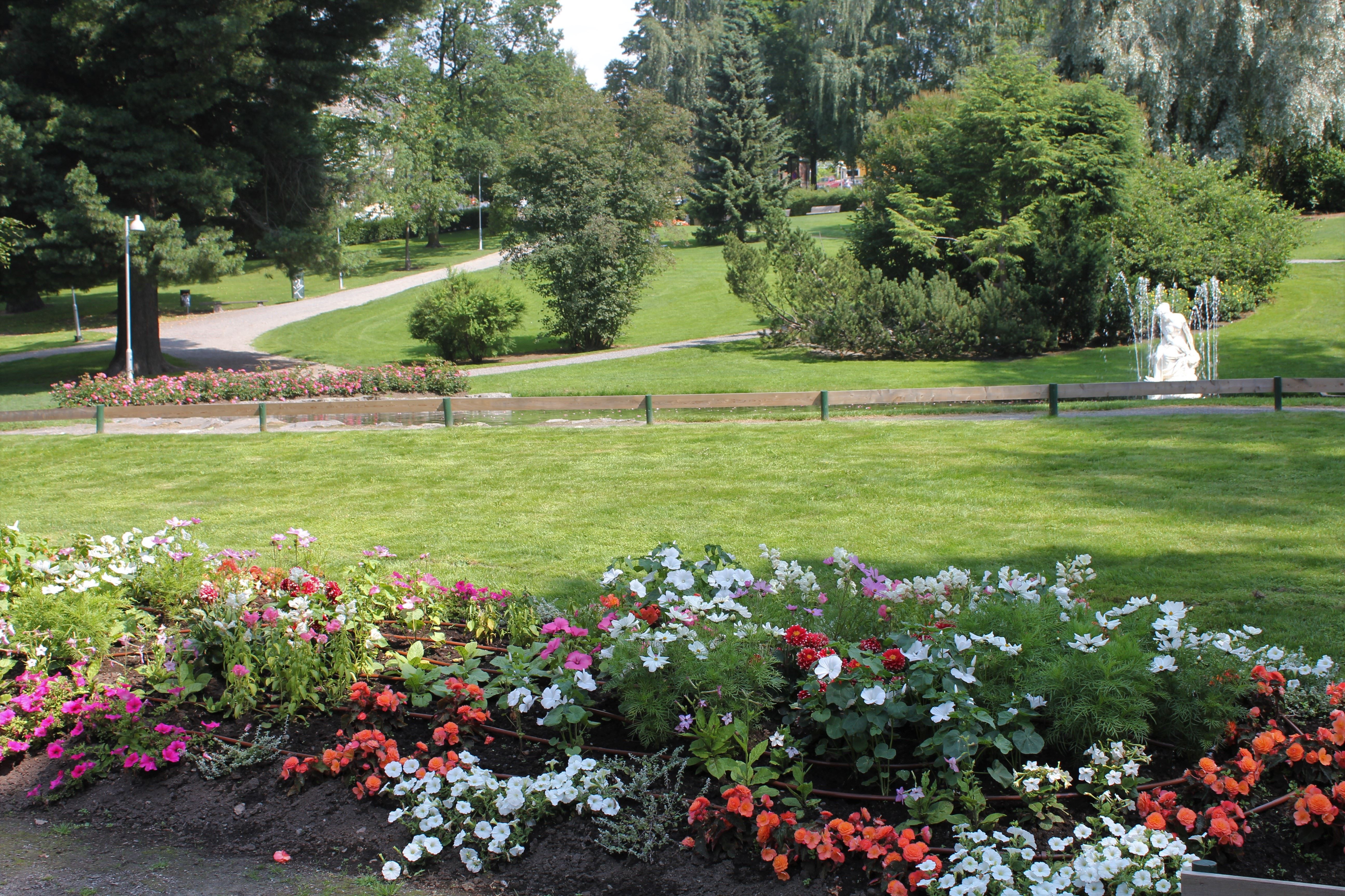 Fargerikt blomsterbed i forgrunnen med grønn plen, gangstier, trær og en fontene med statue i bakgrunnen..
