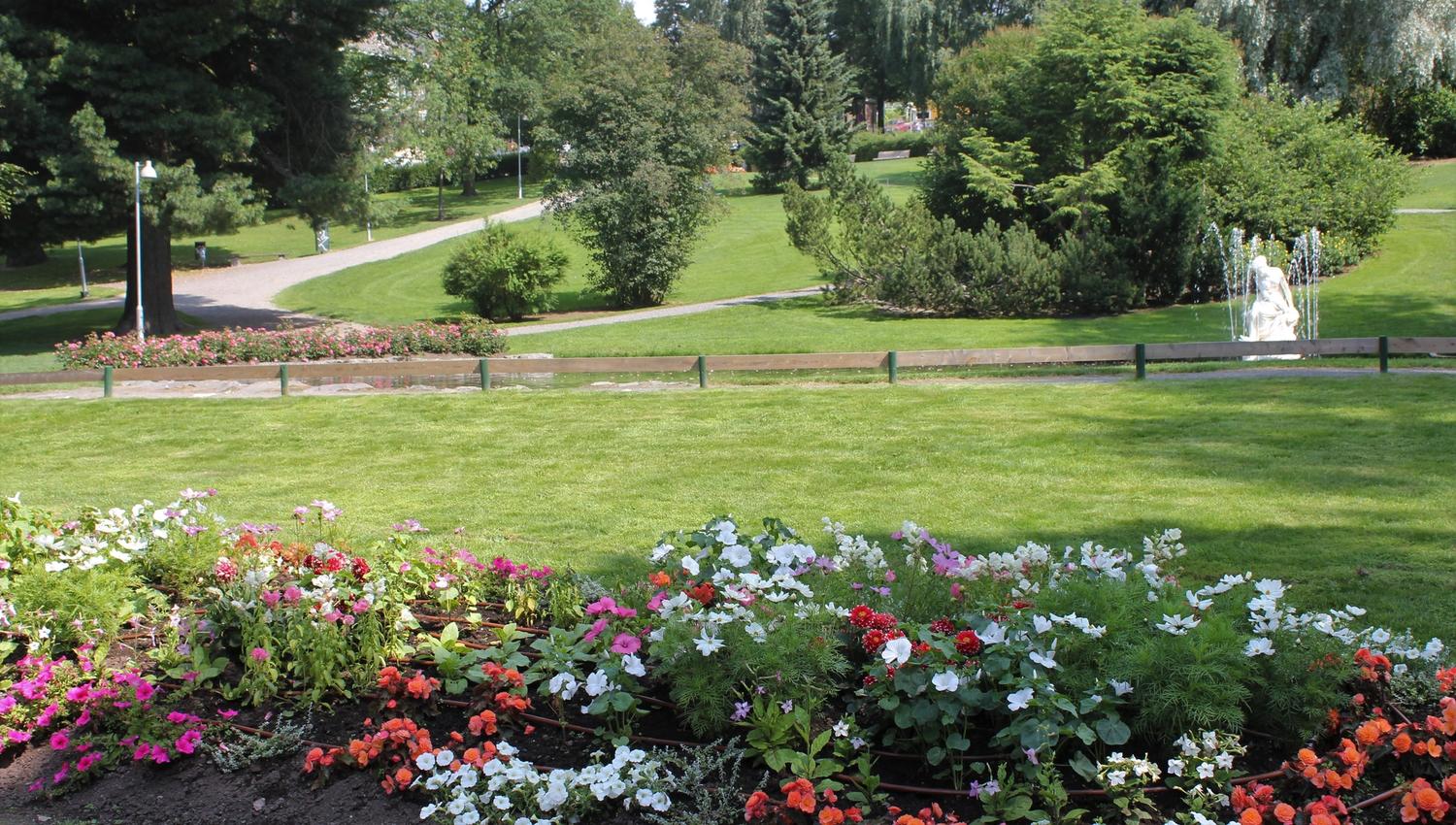Fargerikt blomsterbed i forgrunnen med grønn plen, gangstier, trær og en fontene med statue i bakgrunnen..