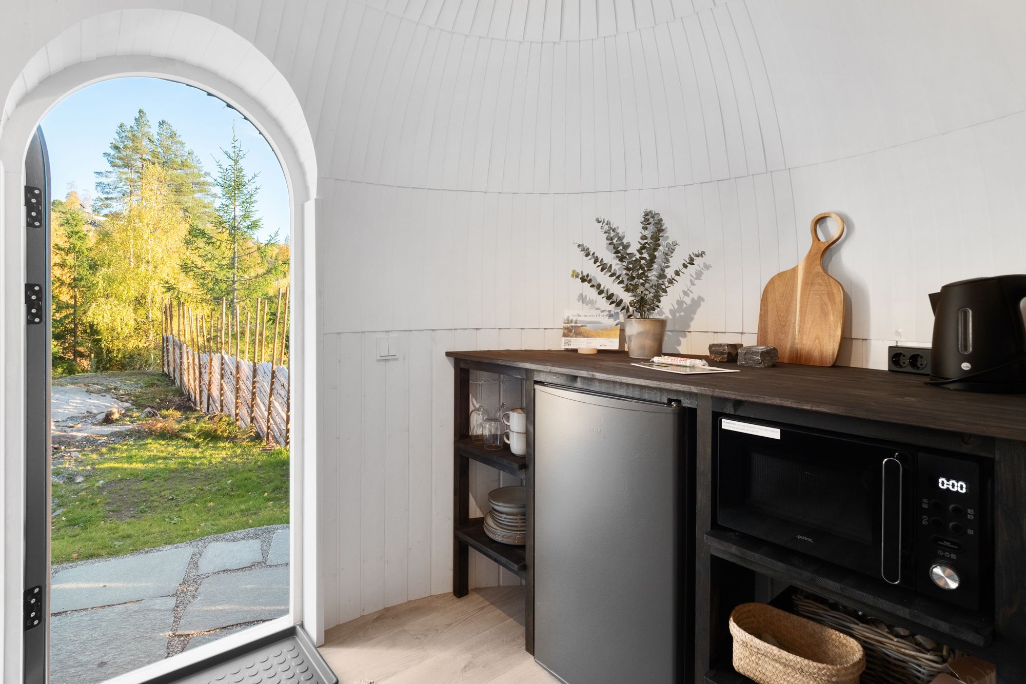 Lite kjøkken med en dør som står åpen med utsikt mot et gjerde og grønn skog