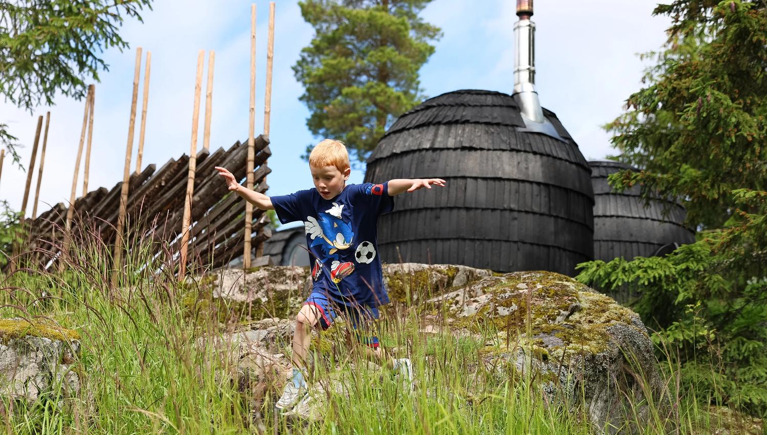 A child playing in the grass near a barrel-shaped grill hut in a forest setting