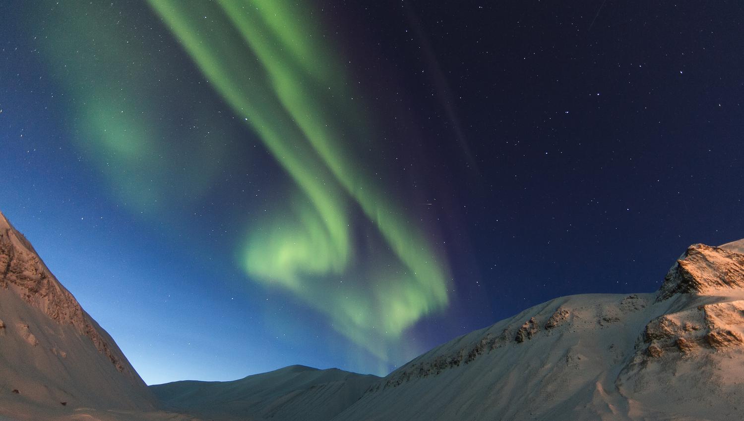 Et fjellandskap i forgrunnen med nordlys på himmelen i bakgrunnen