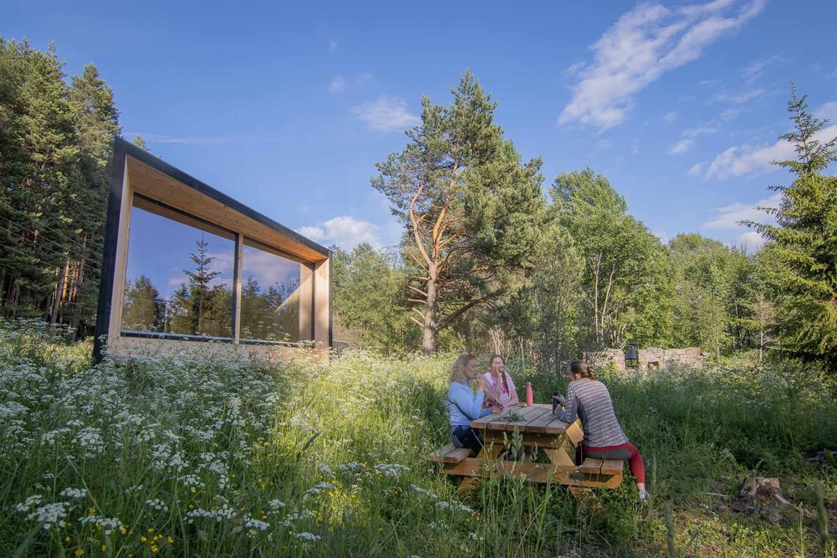 Et lite moderne hyttebygg står i en blomstrende skogeng, der tre personer sitter ved et piknikbord.