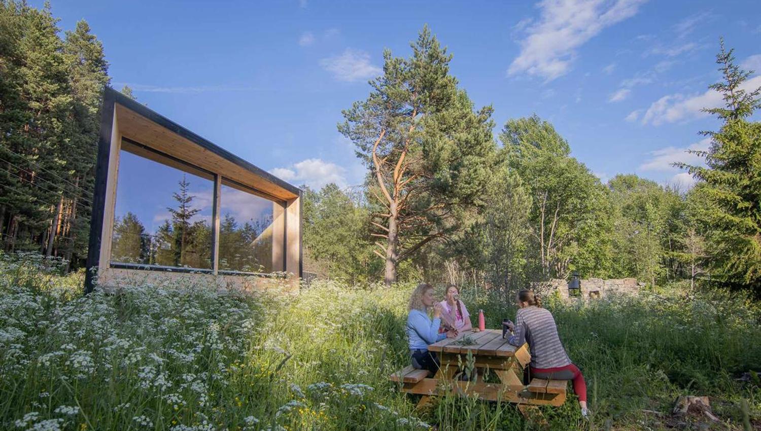 Et lite moderne hyttebygg står i en blomstrende skogeng, der tre personer sitter ved et piknikbord.