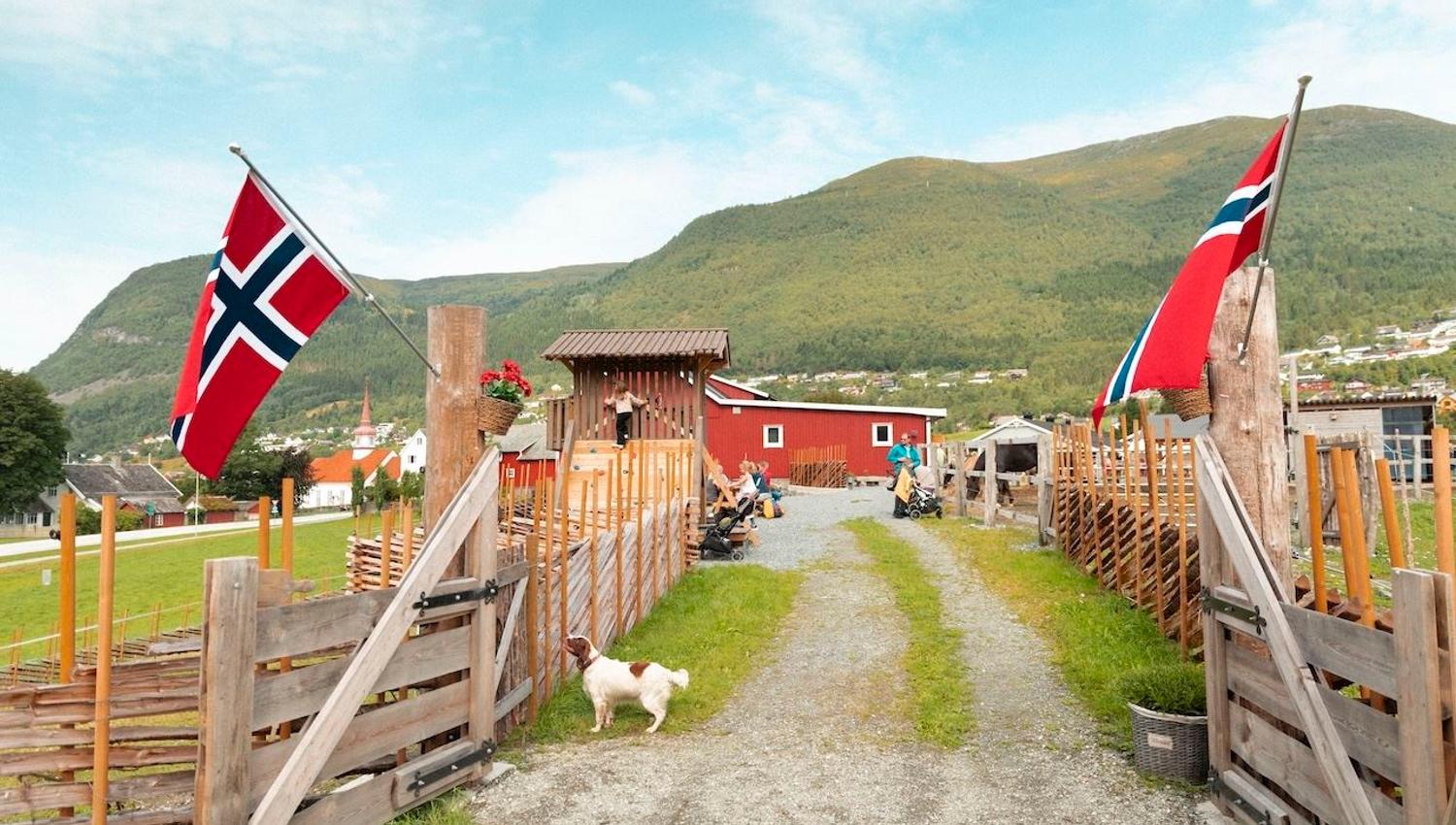 Ein grusveg går gjennom ein gardsport på Myklebust Farm med norske flagg, gjerde og eit lite tun med fjell i bakgrunnen..