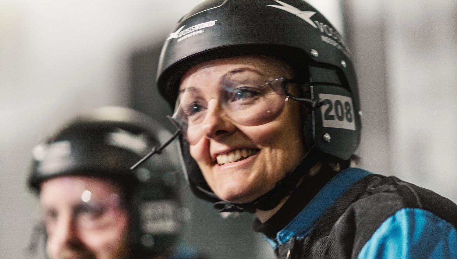 Smiling participant wearing helmet and goggles alongside others, preparing for an indoor flying session.