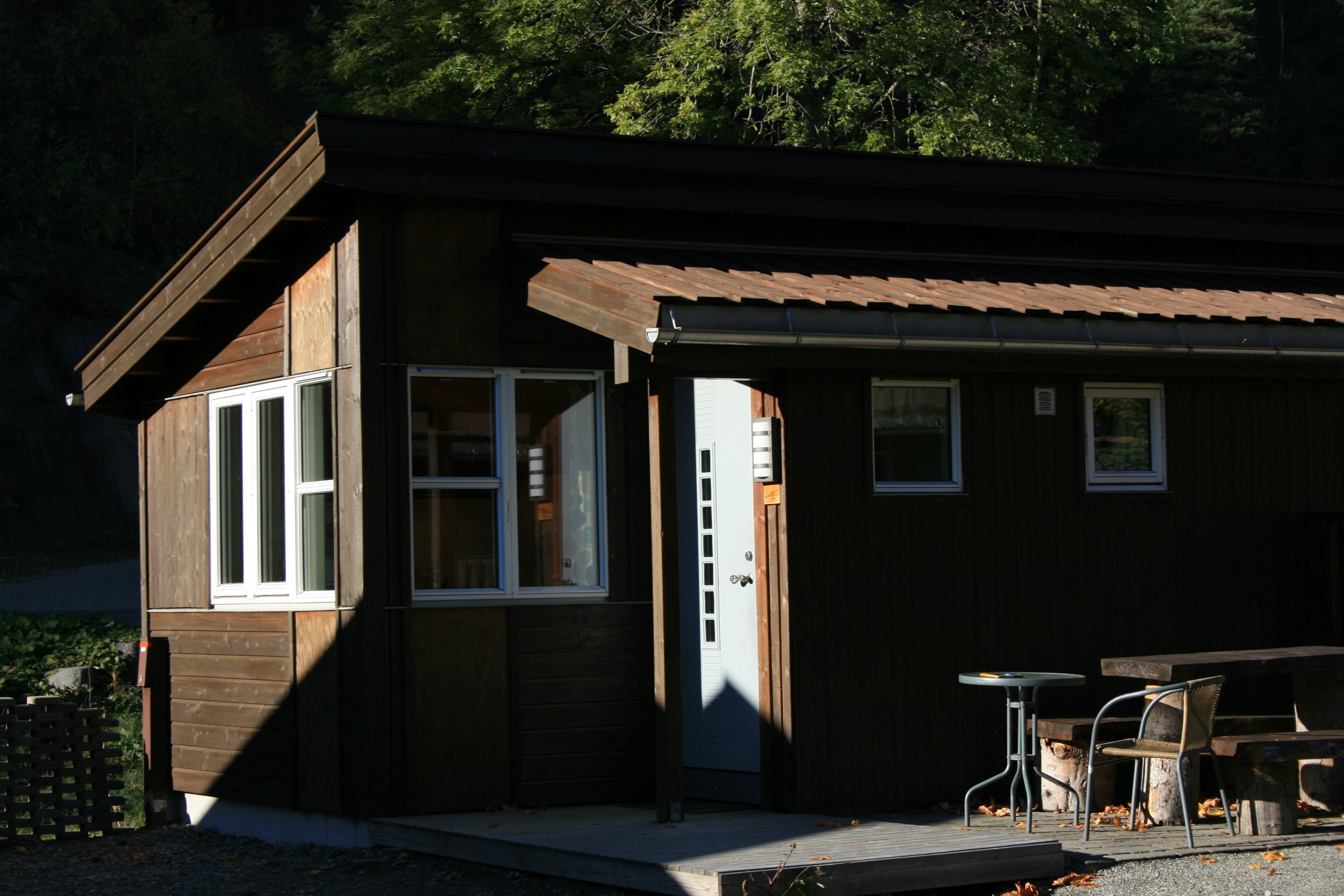 Lita hytte i ettermiddagssol med utebenk og skog rundt.
