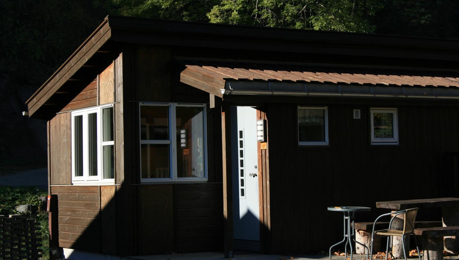 Lita hytte i ettermiddagssol med utebenk og skog rundt.