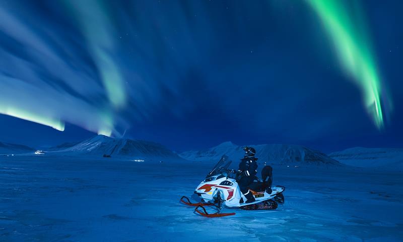 Nordlys over snødekte fjell med en snøscooter stående i front av bildet