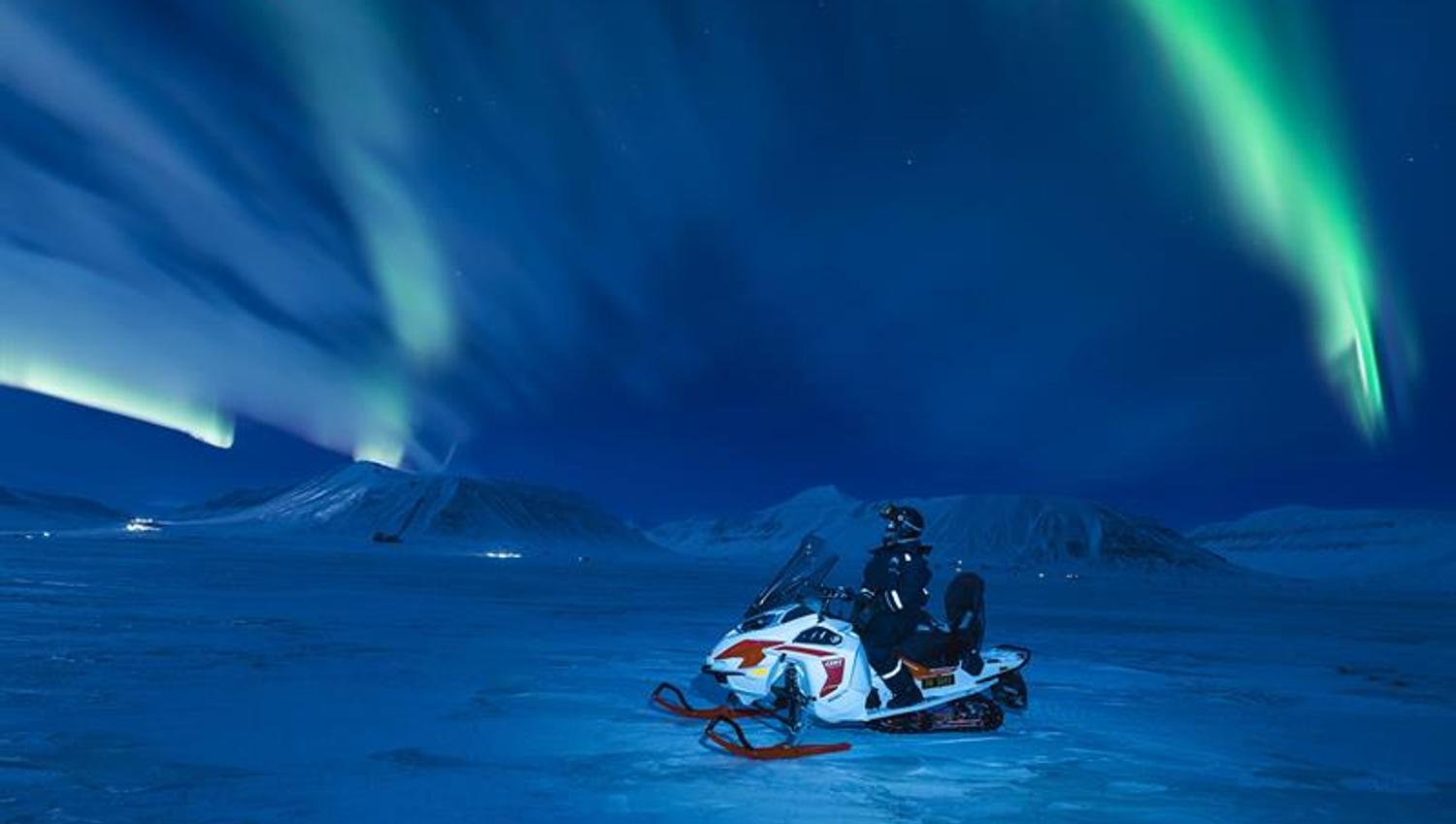 Nordlys over snødekte fjell med en snøscooter stående i front av bildet