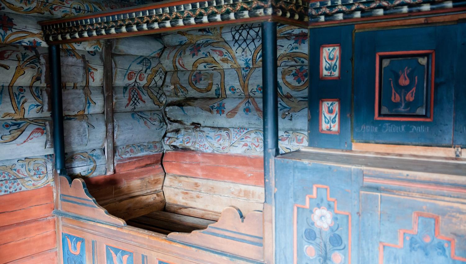 Marnardal Museum og Mjålandsstova, treseng med blå skap vedsidenav. Hvitmalte tømmervegger med fargerike detaljer i rosemaling.