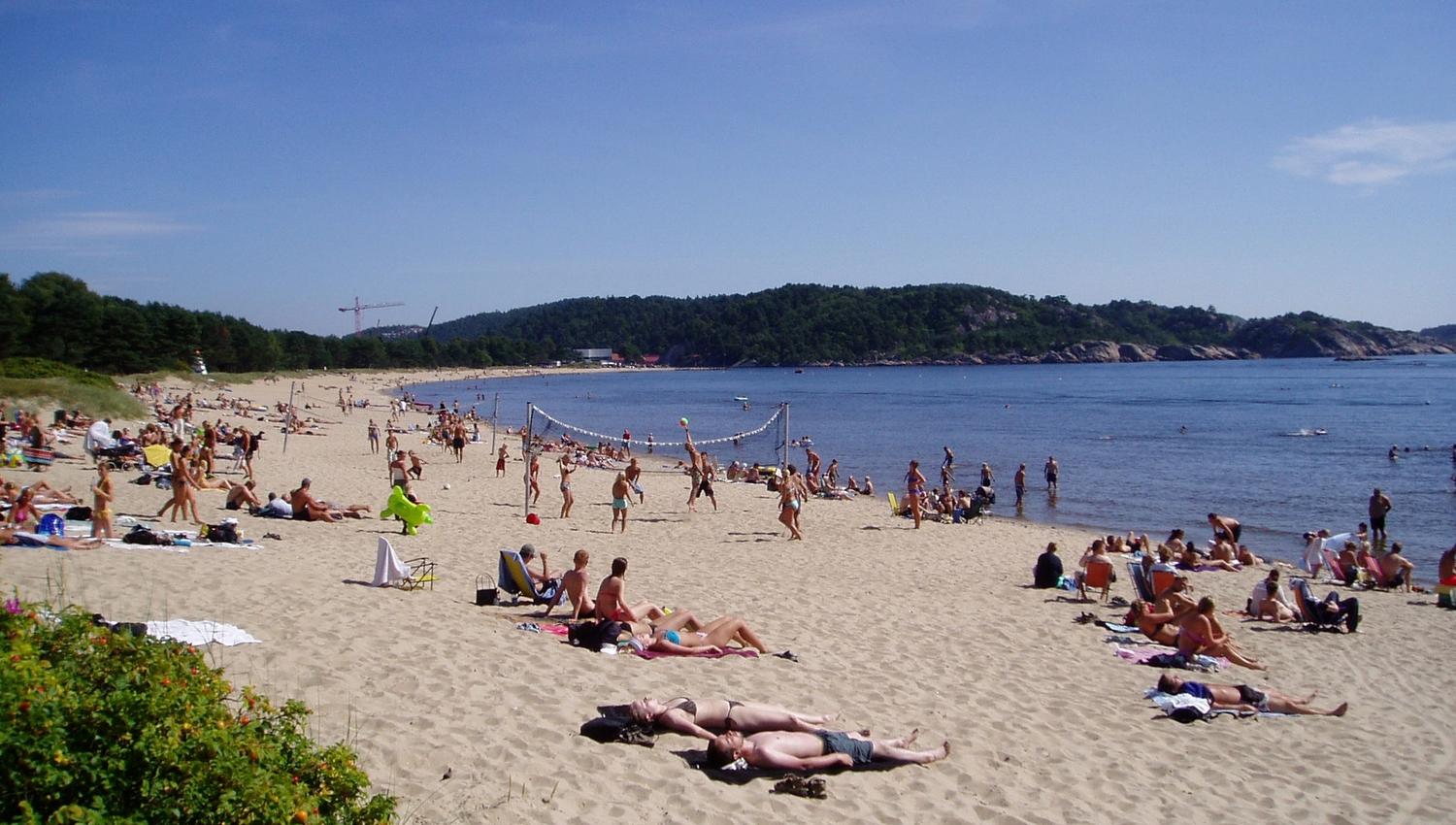 Mange mennesker som soler seg på stranden, mens noen spiller strandvolleyball