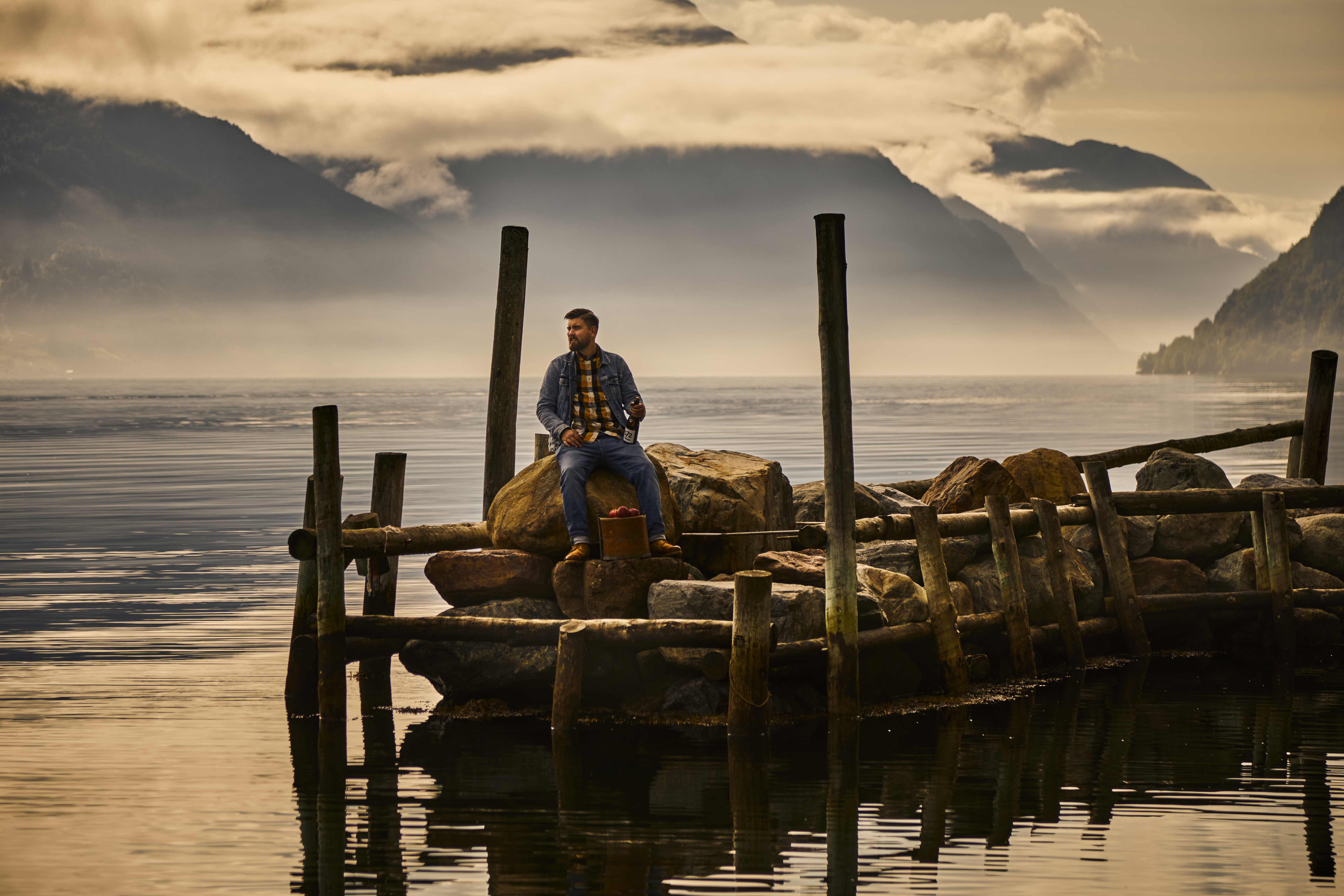 Mann sitter på en brygge med en flaske sider frå Hardanger ved fjordkanten i Sørfjorden i Hardanger.