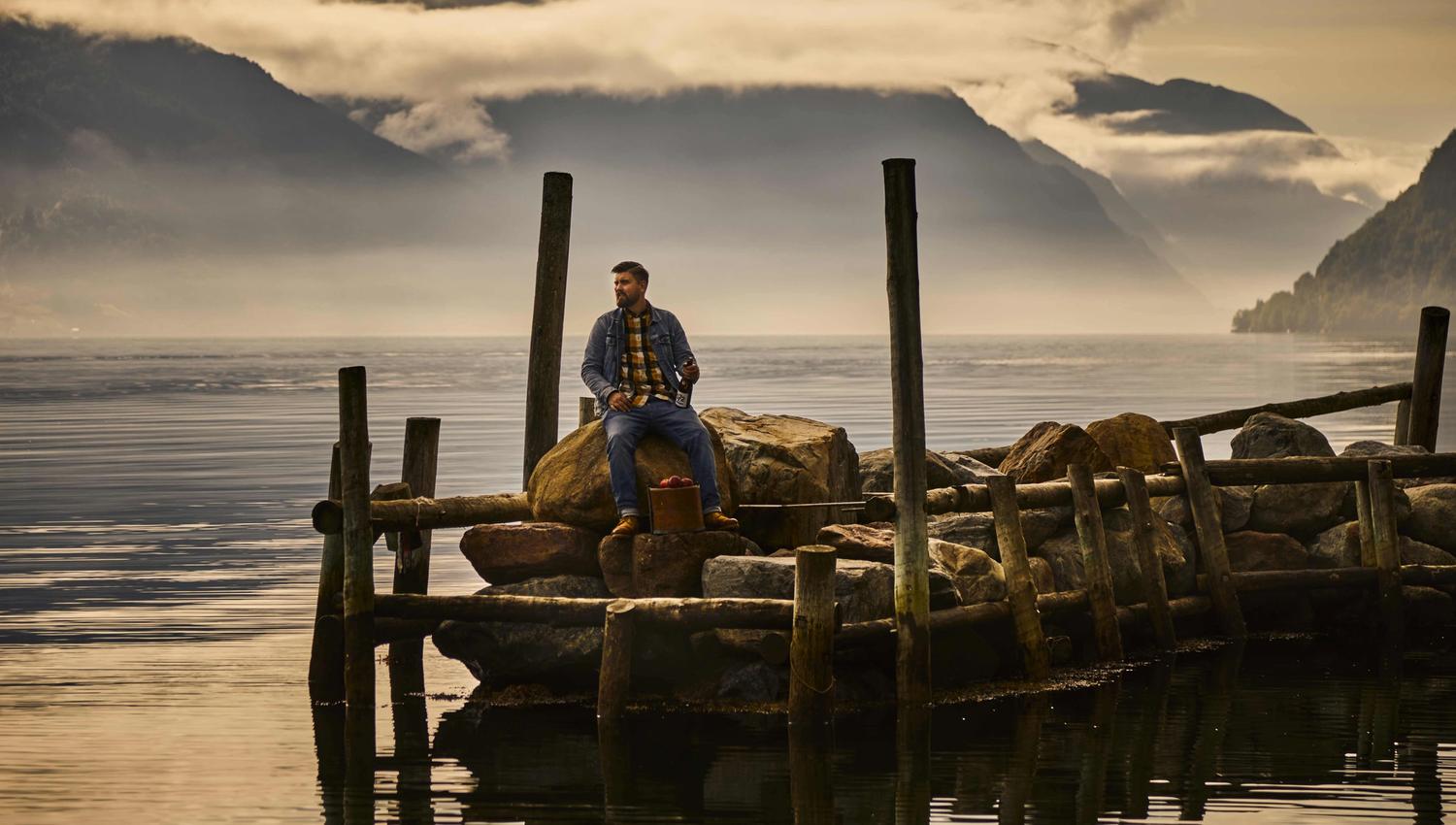 Mann sitter på en brygge med en flaske sider frå Hardanger ved fjordkanten i Sørfjorden i Hardanger.