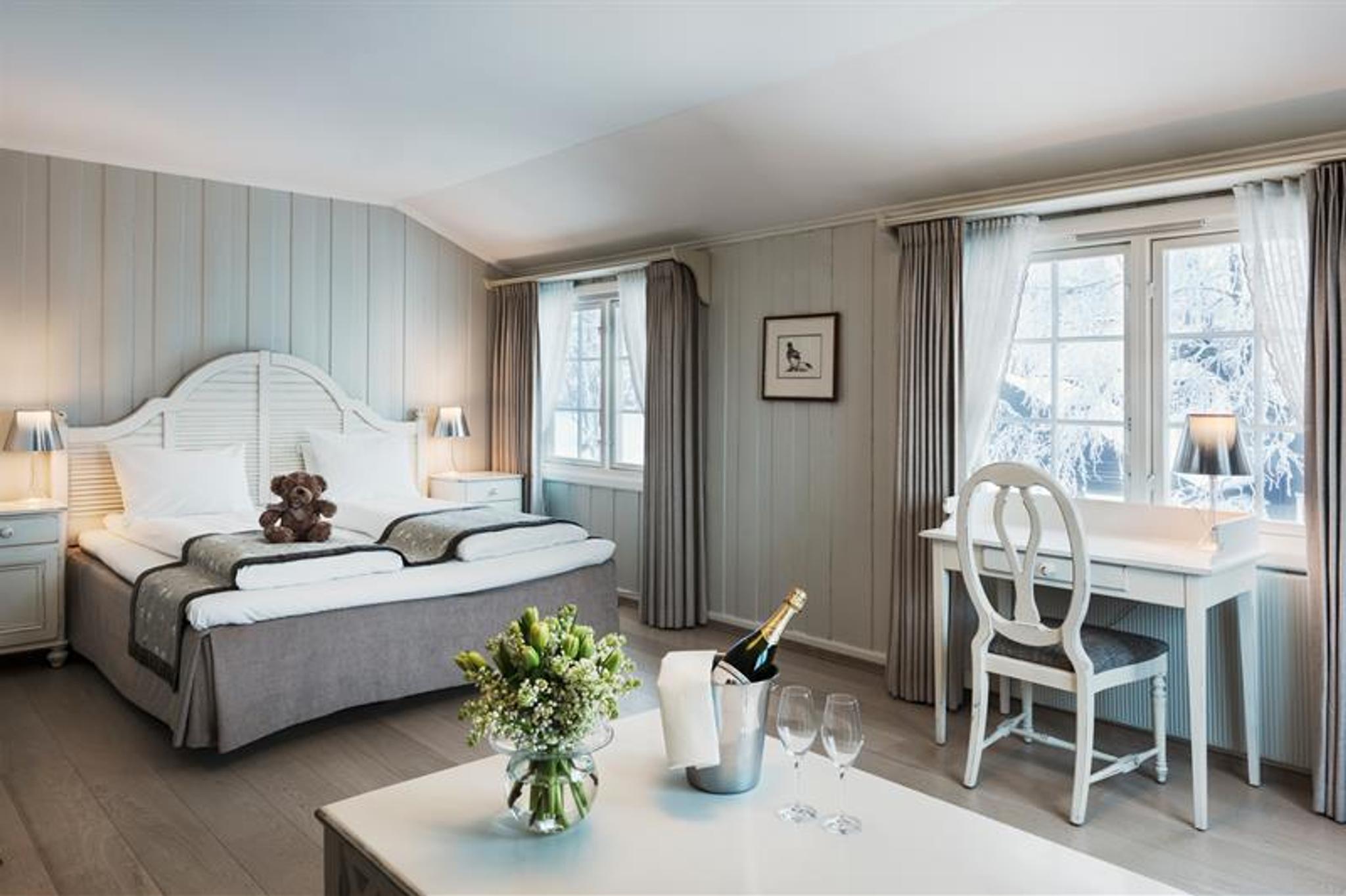 Dobbeltrom med grått og hvitt interiør. Skrivepult, teddybjørn på sengen, champagne og blomster på bordet. Foto