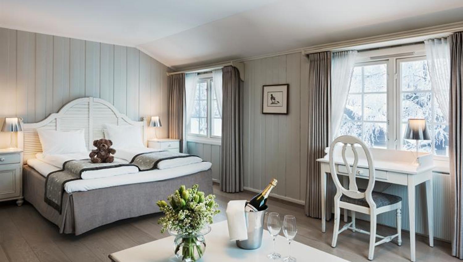 Dobbeltrom med grått og hvitt interiør. Skrivepult, teddybjørn på sengen, champagne og blomster på bordet. Foto