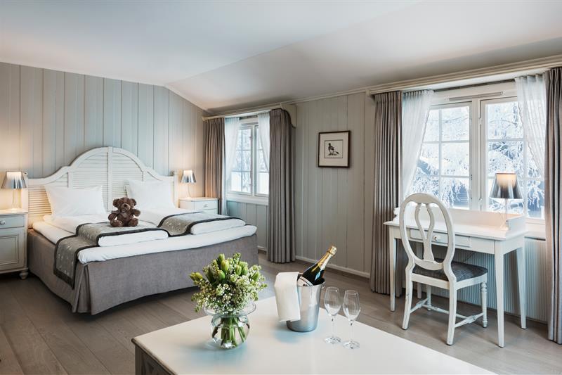 Dobbeltrom med grått og hvitt interiør. Skrivepult, teddybjørn på sengen, champagne og blomster på bordet. Foto