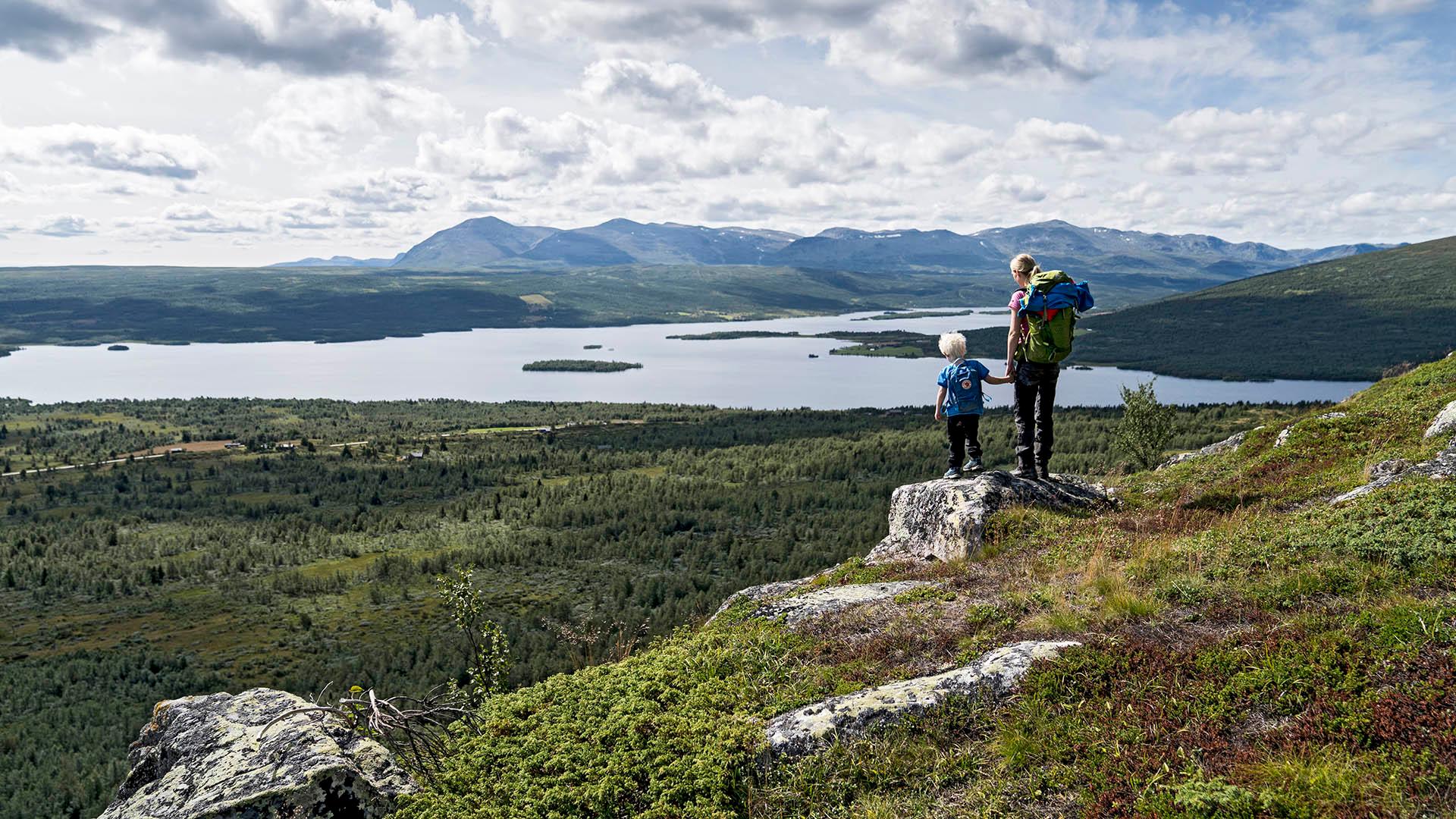 Kvinne og barn står på liten stein og skuer utover et storslått landskap, med innsjø og fjell i bakgrunnen.