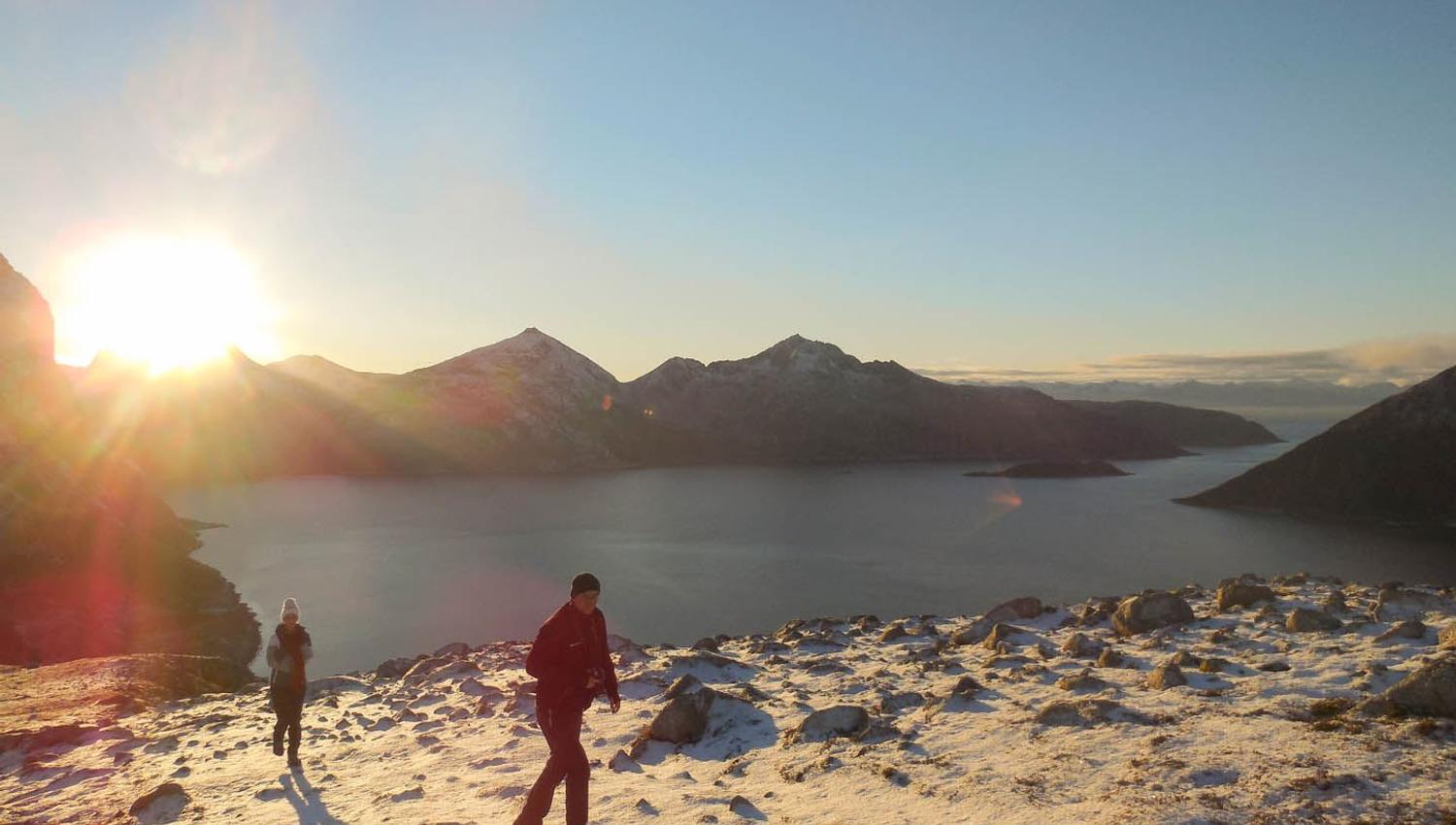 Two people walking in snowy mountain terrain with sunset and water in the background.