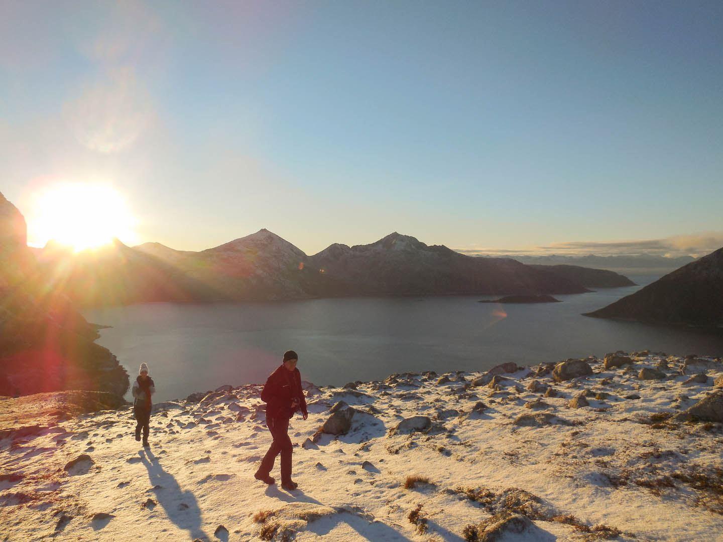 Two people walking in snowy mountain terrain with sunset and water in the background.
