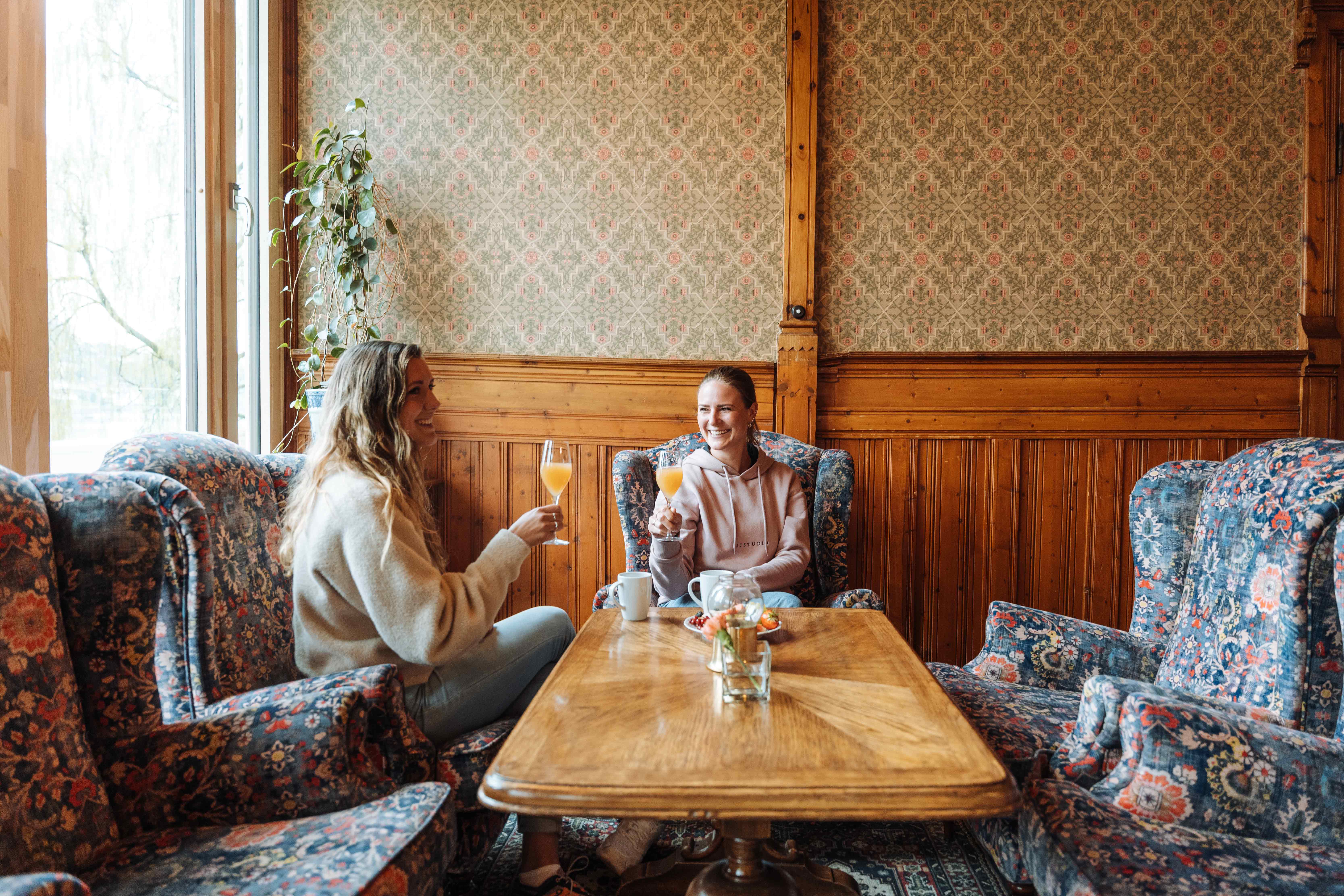 To venninner sit i ei sjarmerande stove på Thon Hotel Sandven og nyt eit glas fersk juice, omgitt av tradisjonell norsk interiørstil.