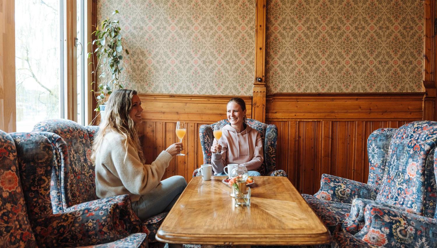 To venninner sit i ei sjarmerande stove på Thon Hotel Sandven og nyt eit glas fersk juice, omgitt av tradisjonell norsk interiørstil.