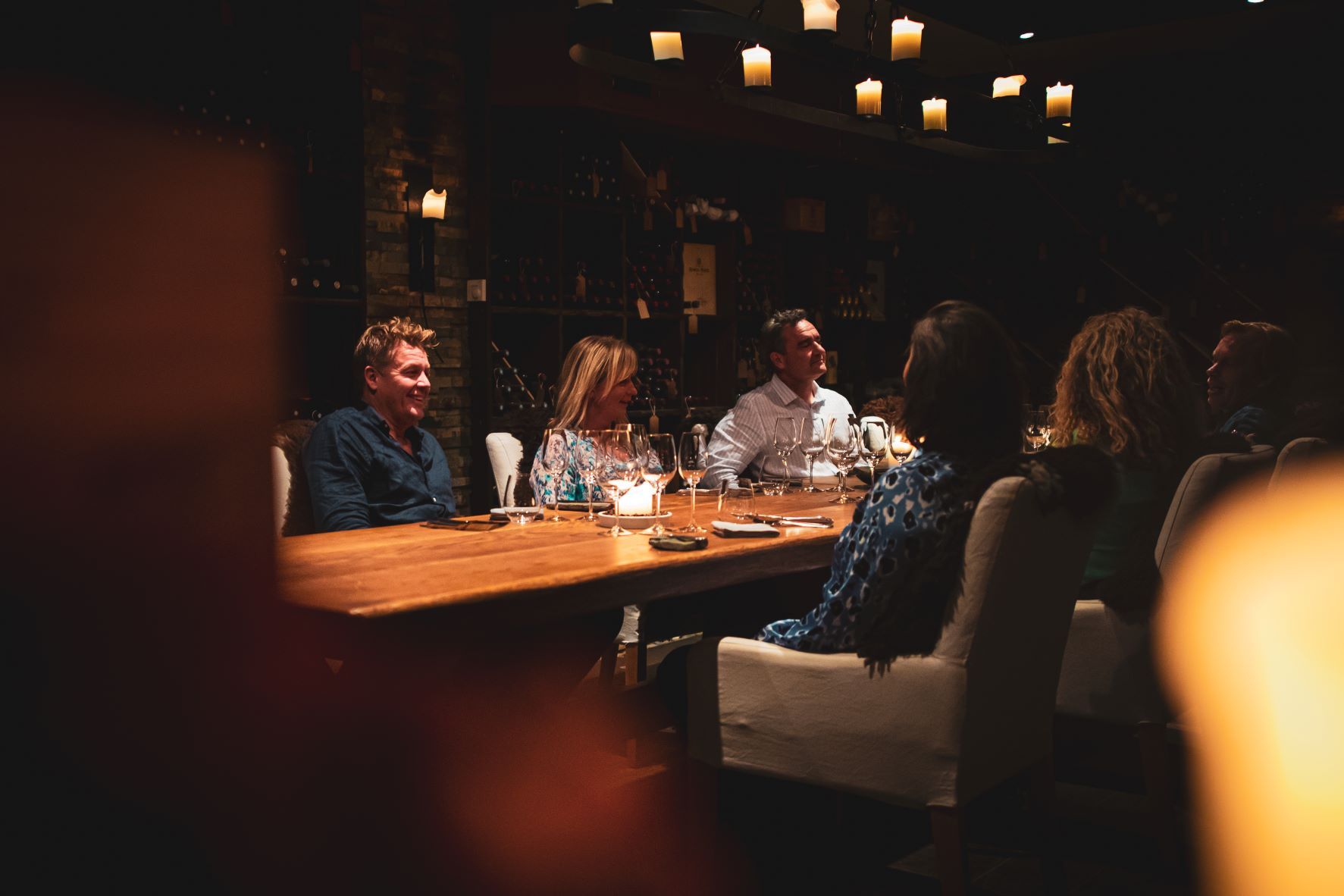 En gruppe personer som sitter langs et langbord med vinglass på bordet og vinflasker i hyller i bakgrunnen