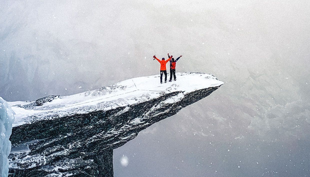 Two winter hikers standing on Trolltunga in light snowfall with expansive mountain views.