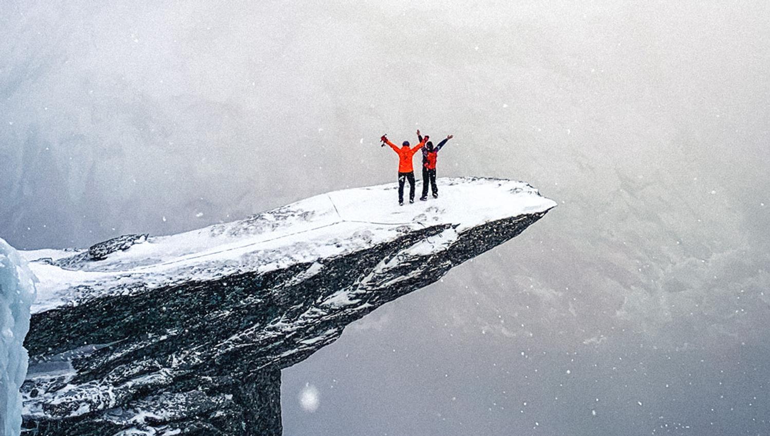 Two winter hikers standing on Trolltunga in light snowfall with expansive mountain views.